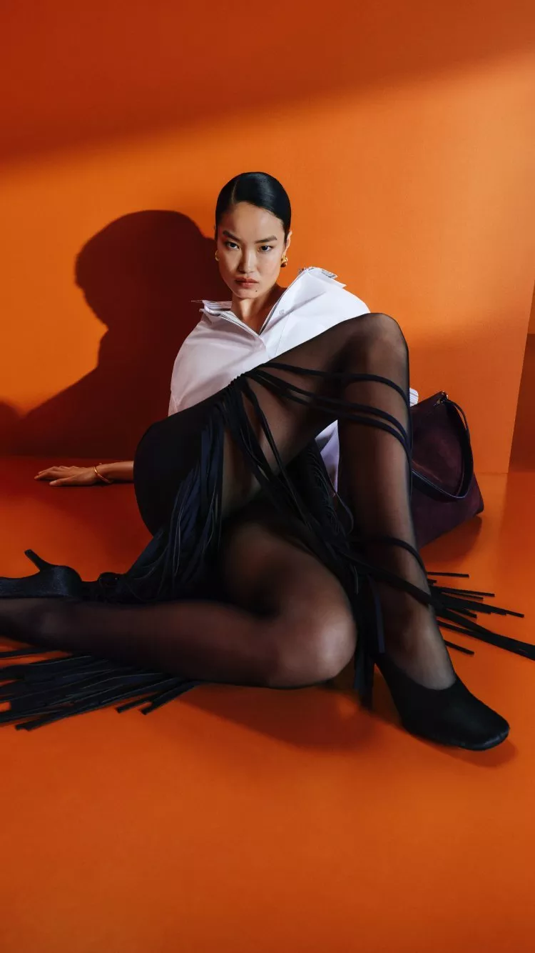 Fashion model in white shirt and black sheer tights posing on orange studio set with handbag and heels
