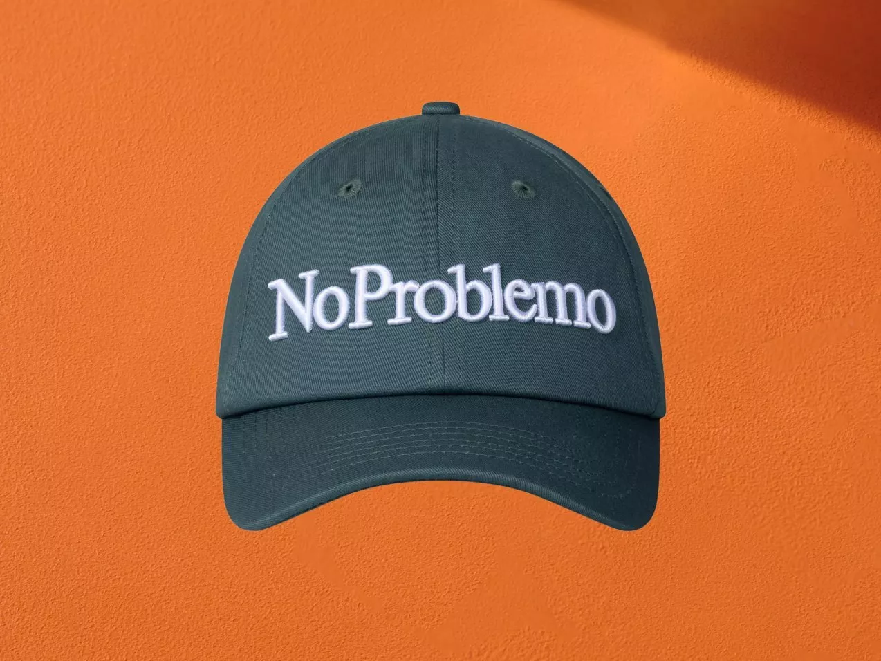 Dark green baseball cap with white “NoProblemo” embroidery, front view on orange background, casual streetwear hat