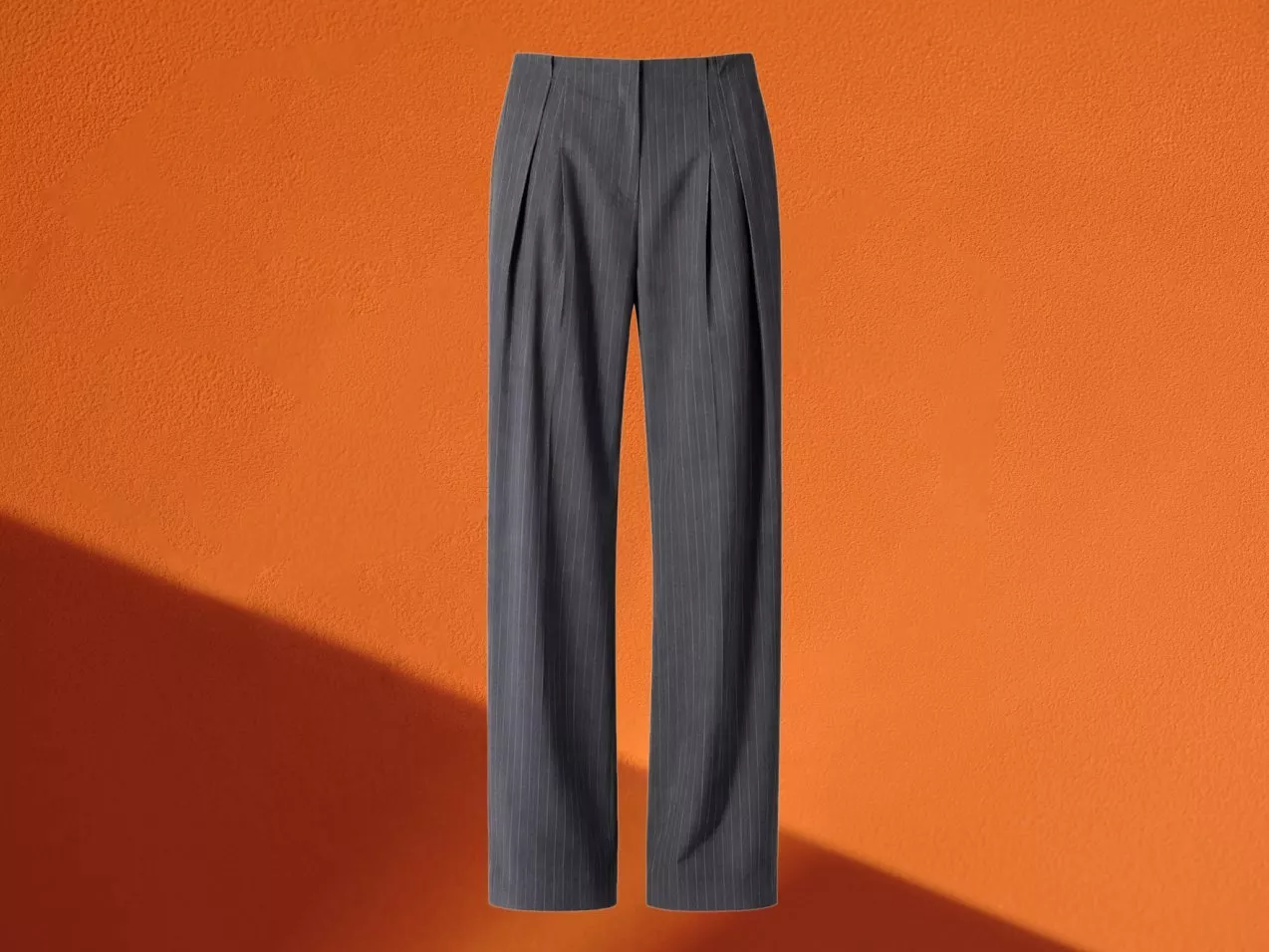 Charcoal grey pinstripe tailored trousers with pleated front, wide-leg fit, on an orange textured background