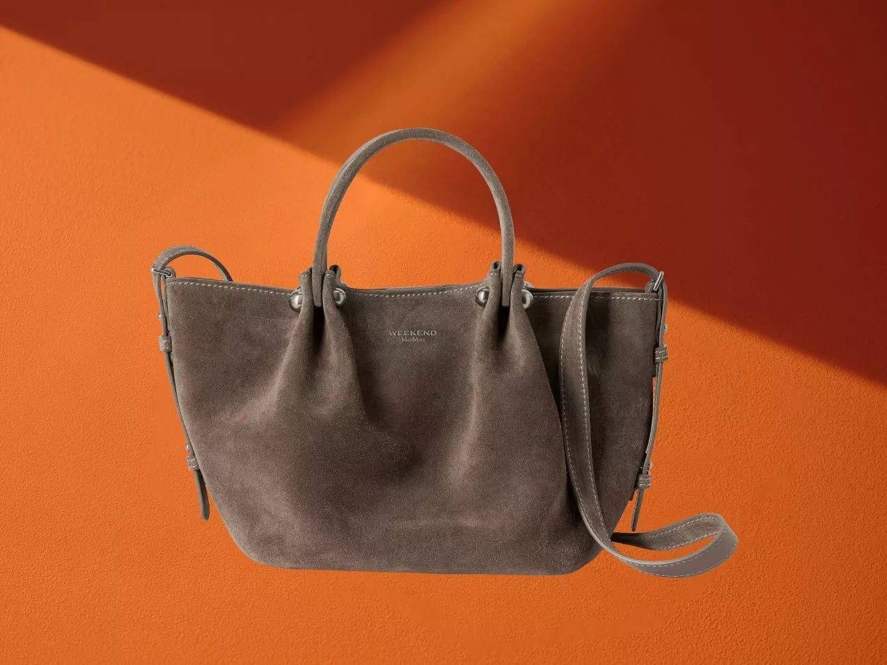 Grey suede tote handbag with top handles and shoulder strap on an orange background, studio product photo