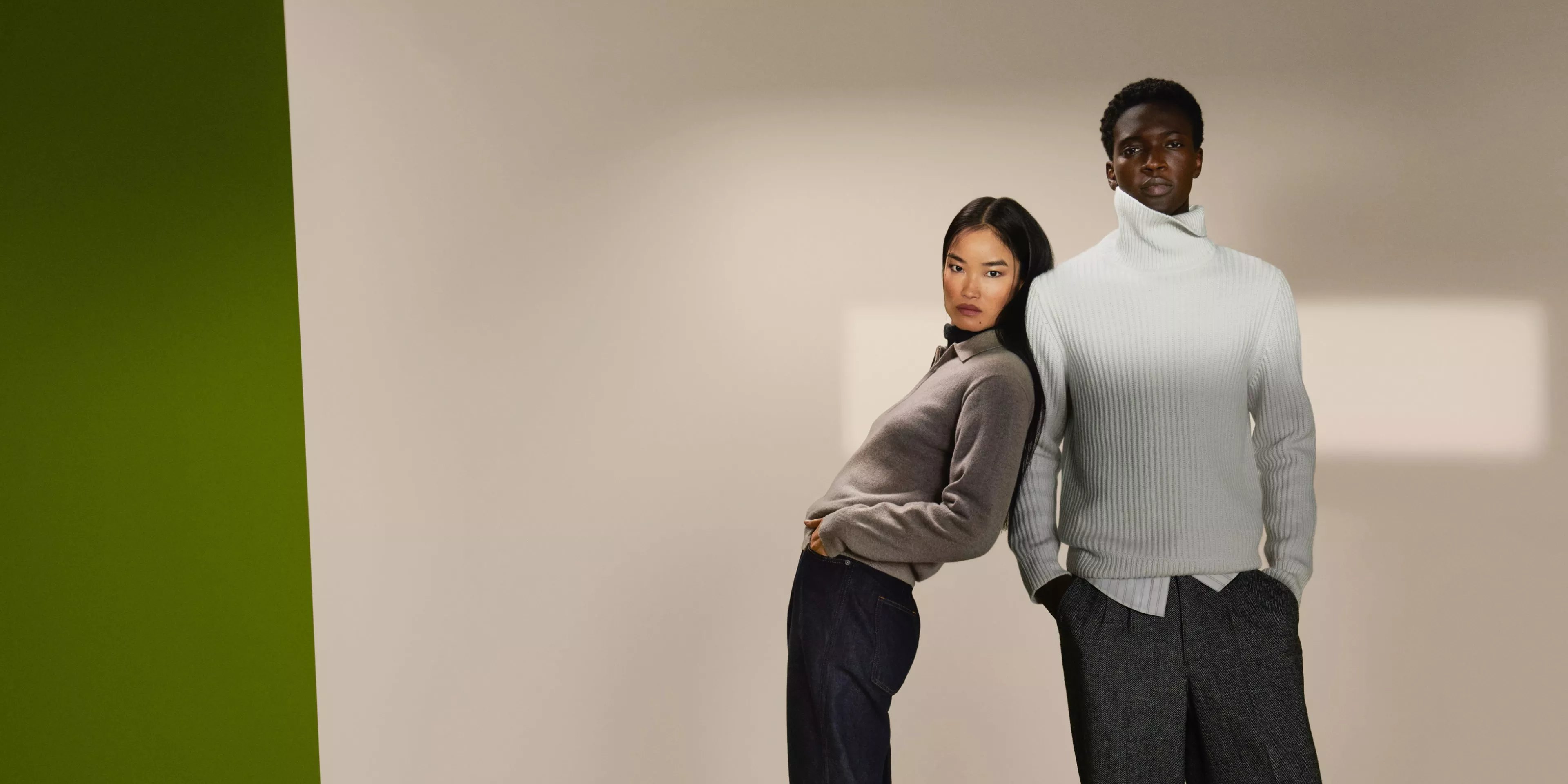 Two fashion models in knitwear, woman in grey jumper and man in white roll neck, studio background with green panel