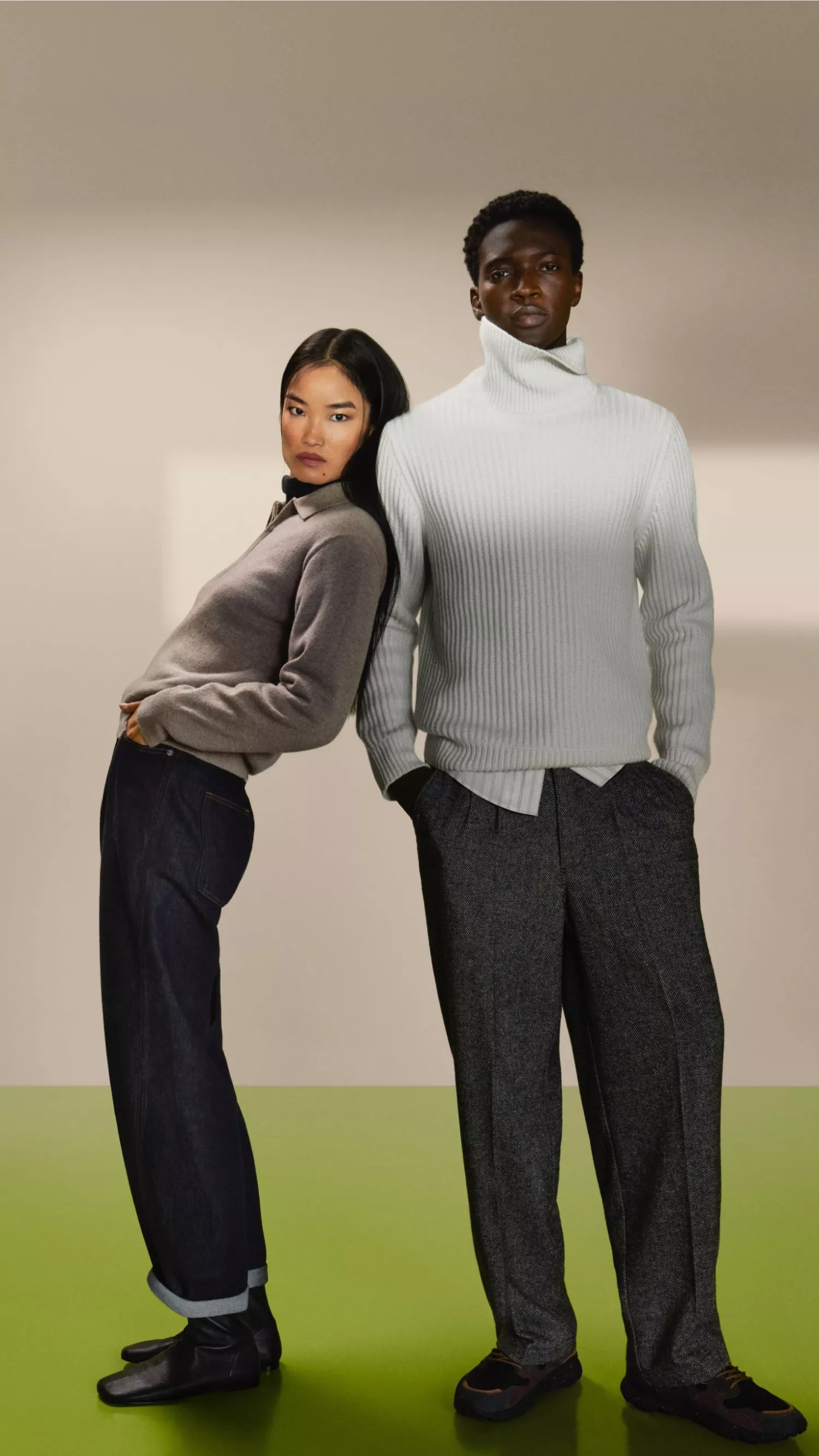Two fashion models in knitwear and trousers posing back-to-back in a studio, showcasing modern winter style.