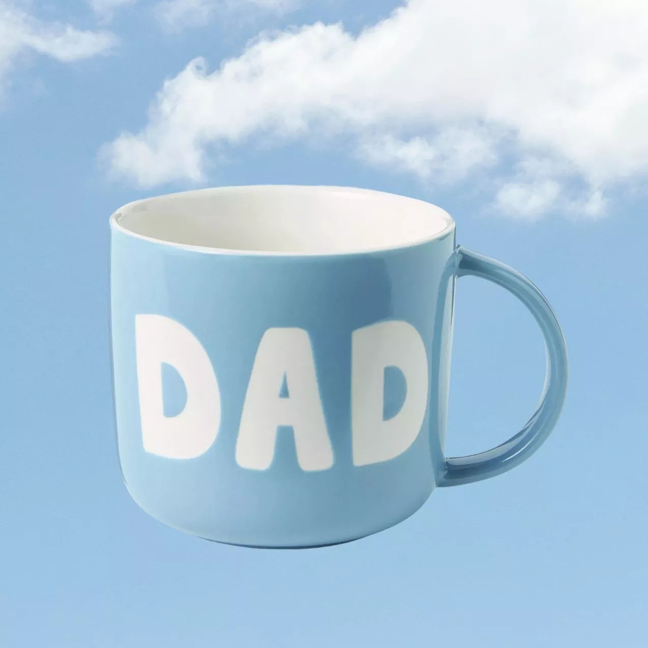 Light blue ceramic mug with bold “DAD” lettering, floating against a blue sky background with soft white clouds