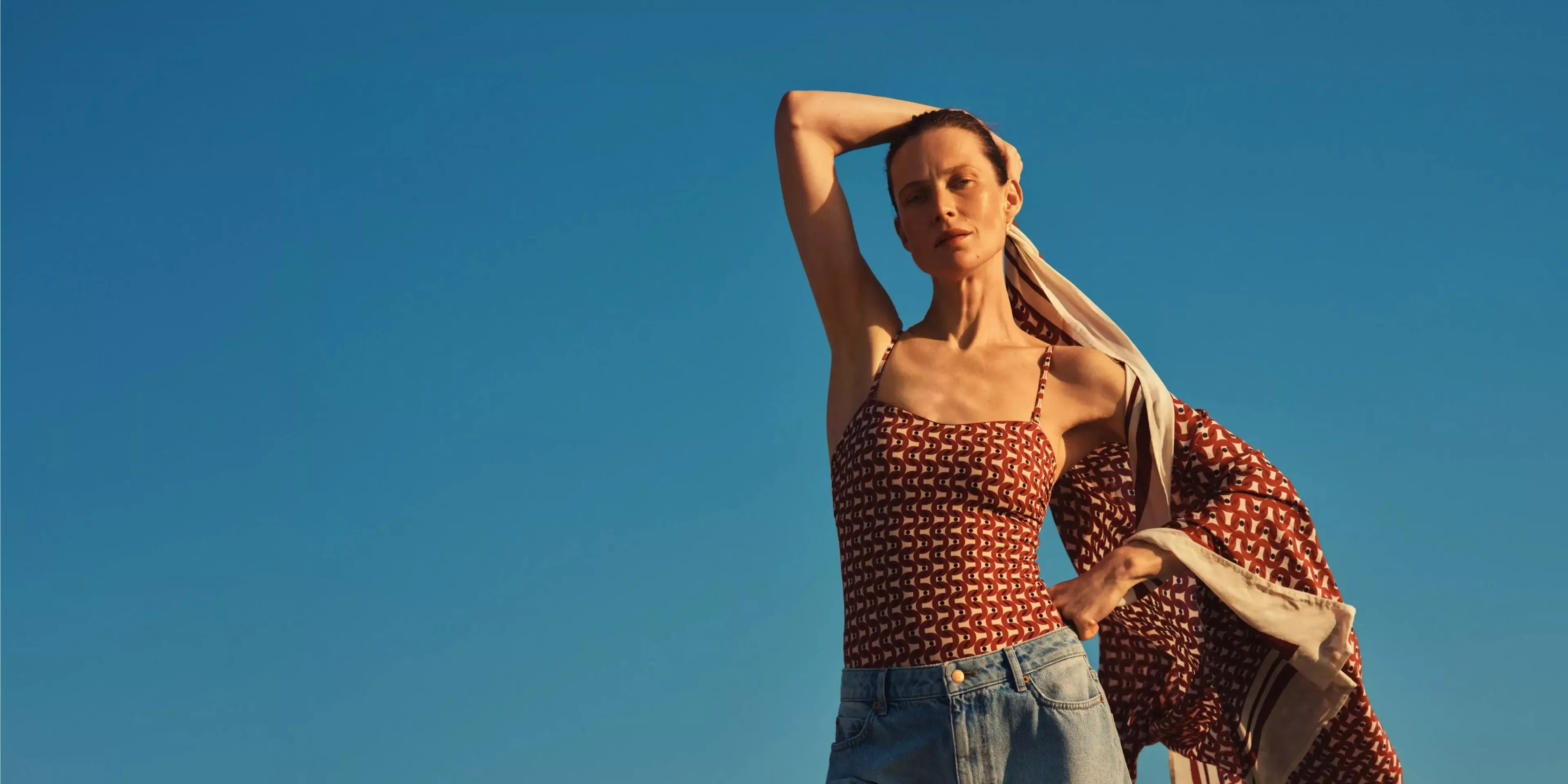 Fashion model in patterned camisole and denim shorts with flowing scarf, posing against a clear blue sky.