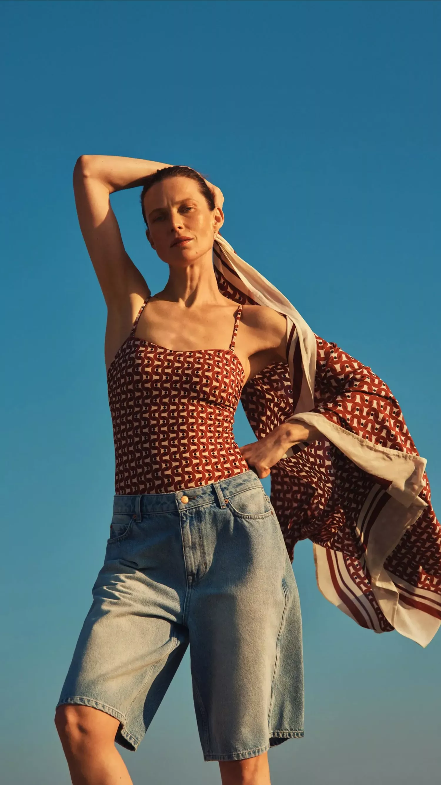 Fashion model in patterned camisole and denim shorts with flowing scarf, posing against a clear blue sky.