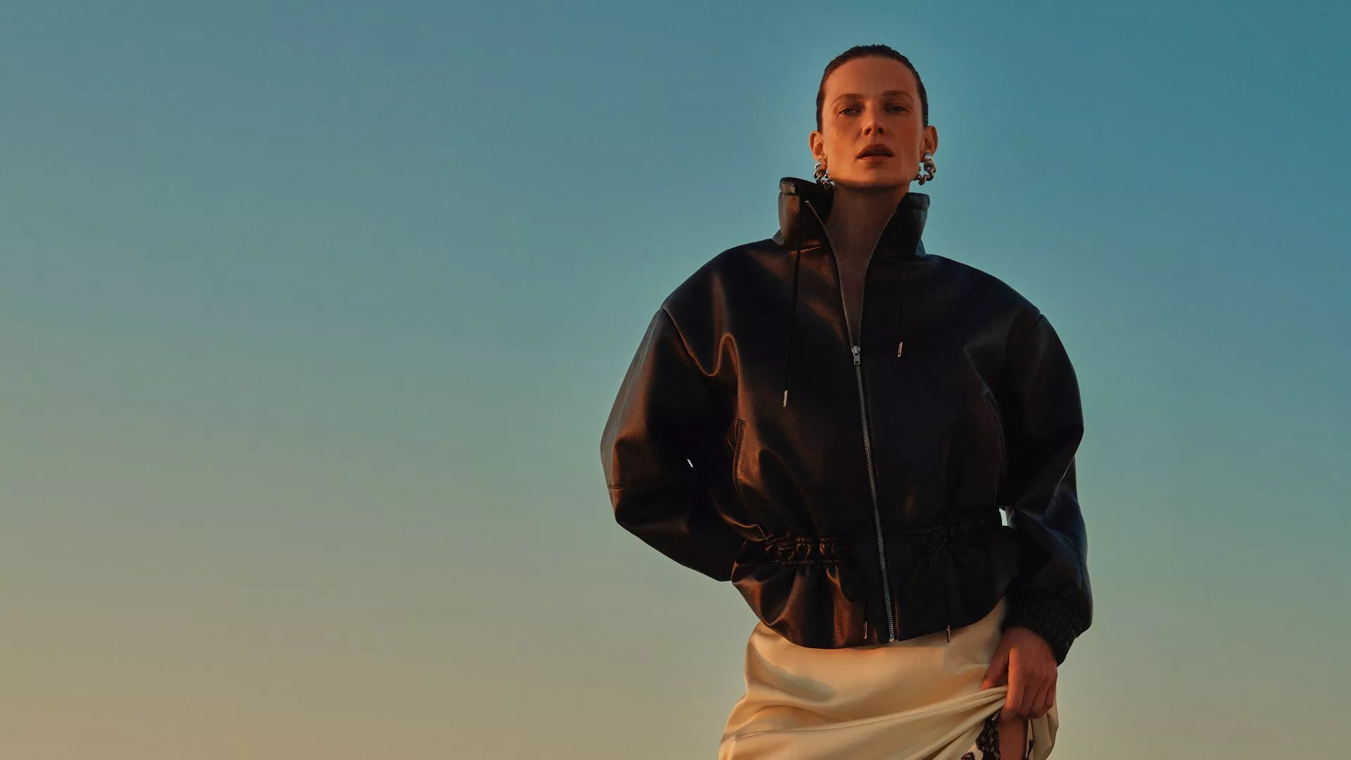 Fashion model in black leather bomber jacket and cream skirt, posing outdoors against a blue sky backdrop