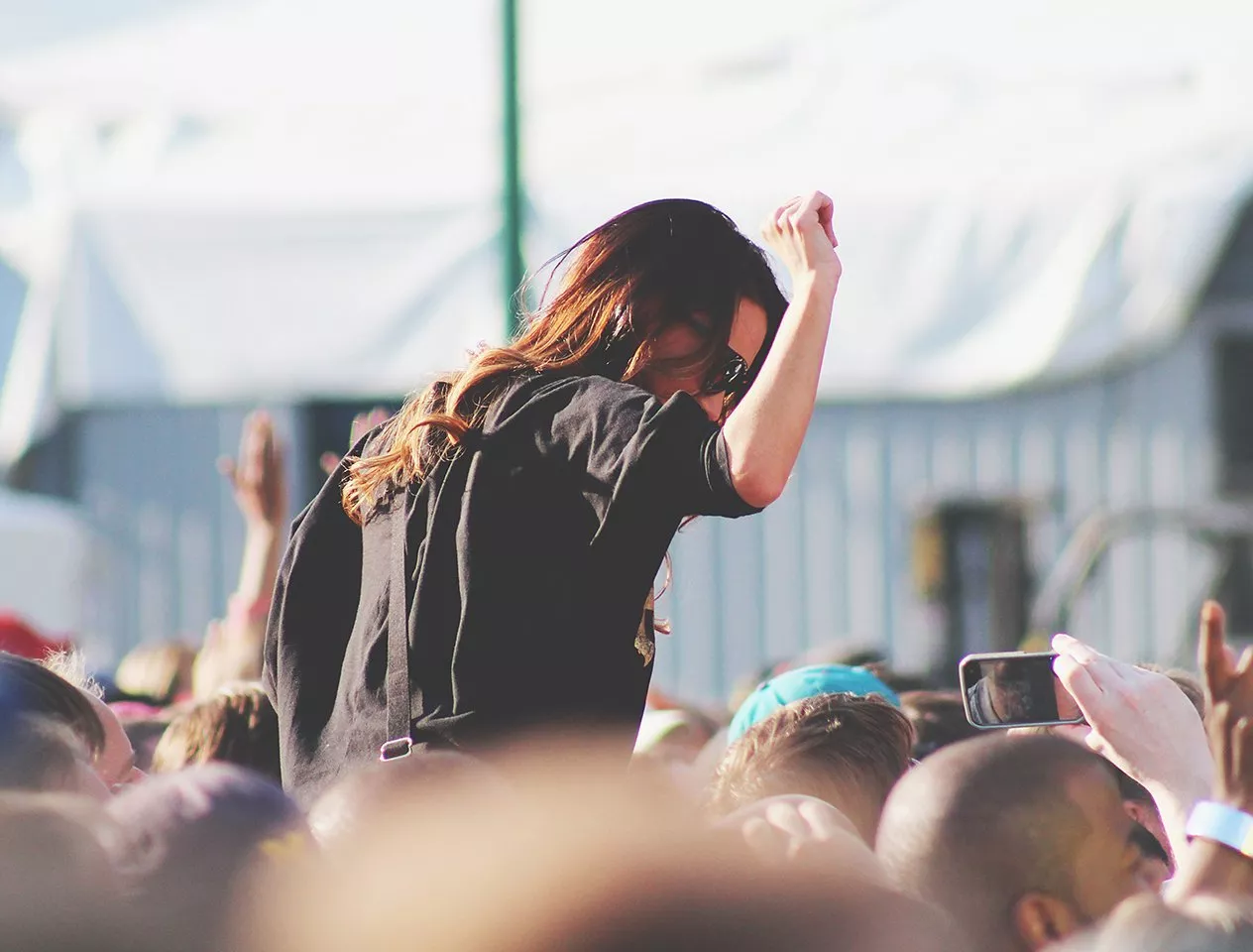 Woman crowd surfing at an outdoor music festival, arm raised, surrounded by cheering fans and phones recording