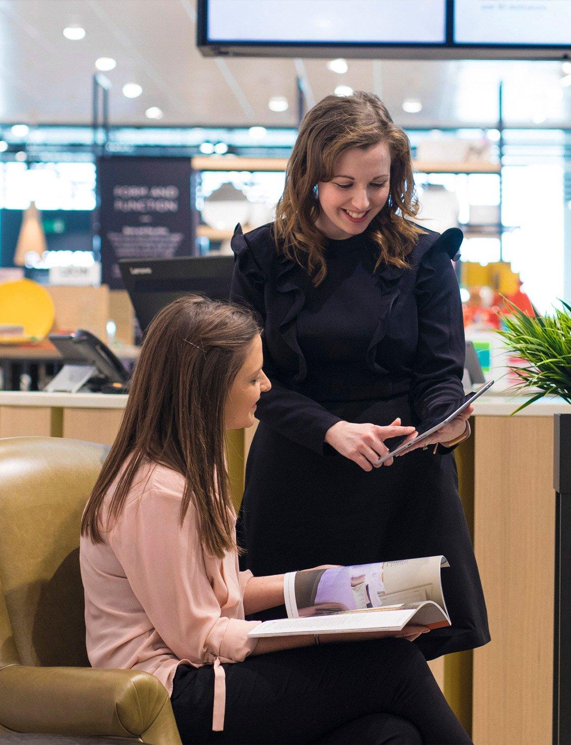 One woman standing showing a tablet to another women sitting in a chair with a brochure in her hand.