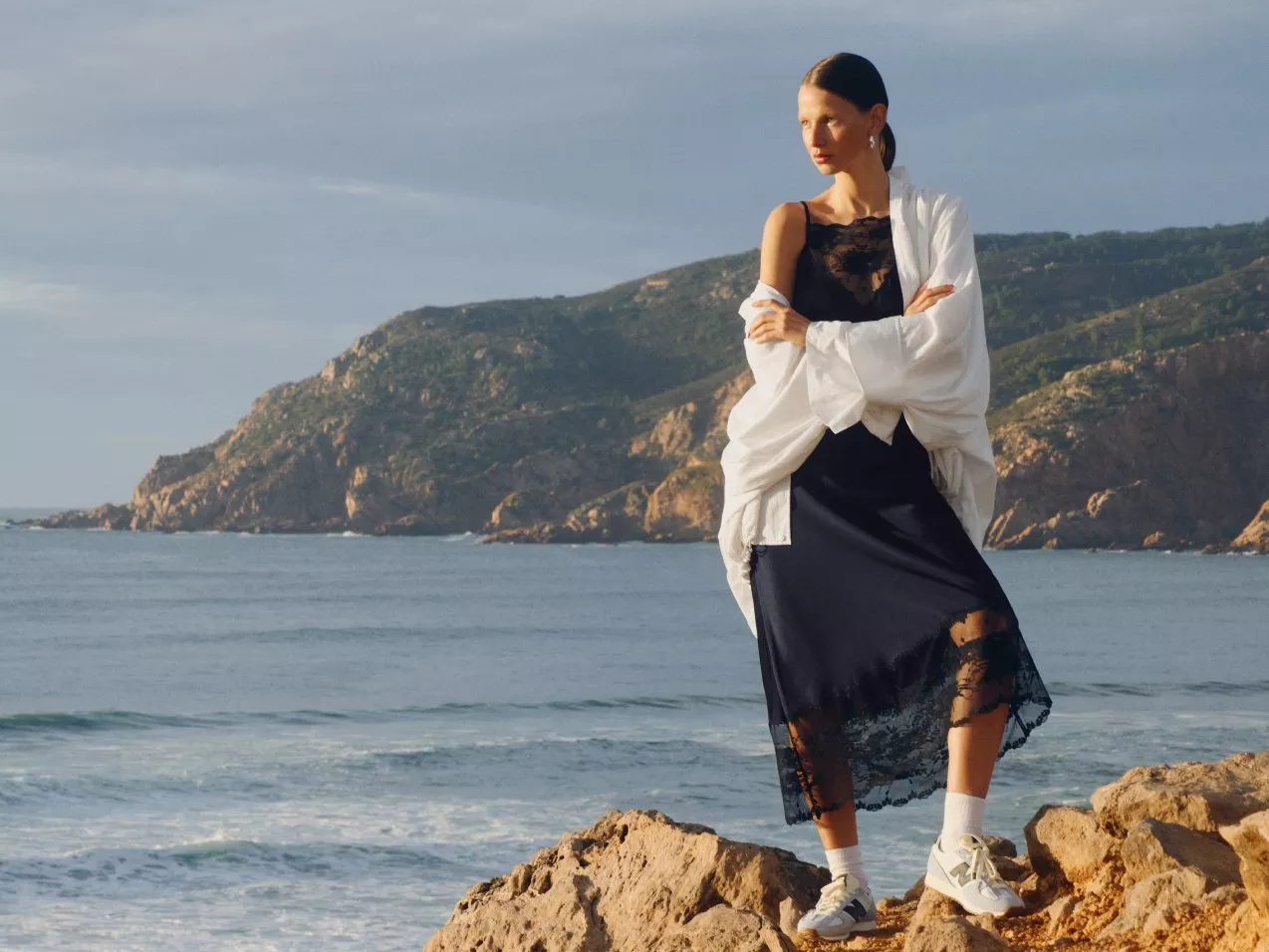 A person stands on rocky cliffs by the sea, wearing a stylish black dress, white trainers, and a flowing white shawl.