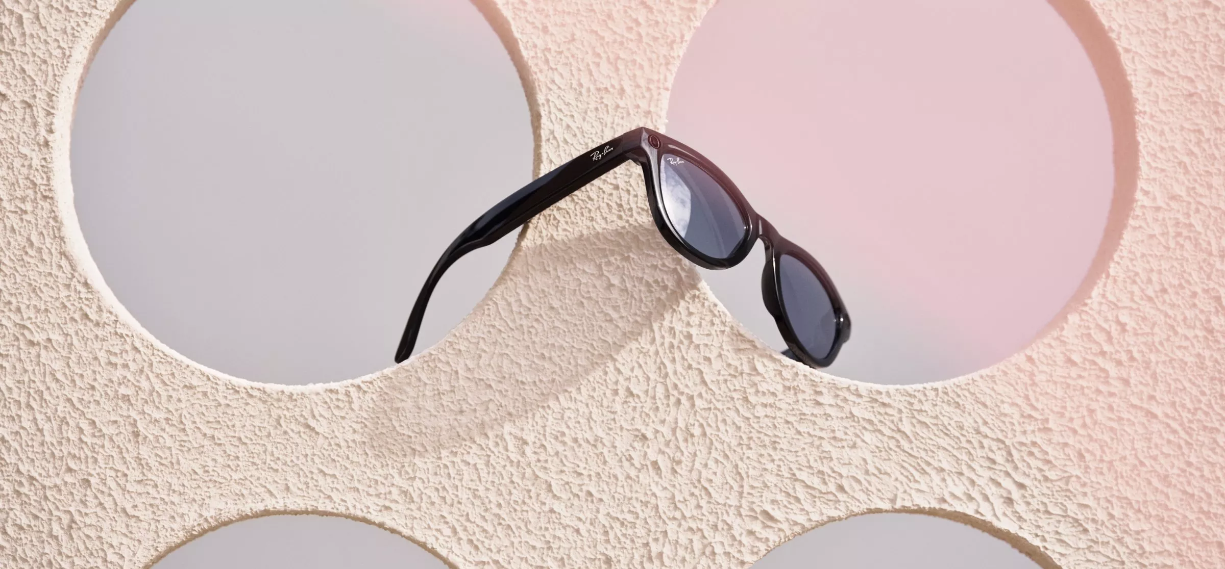 Black Ray Ban Meta sunglasses resting on a textured beige surface with circular cutouts and a soft pink gradient.