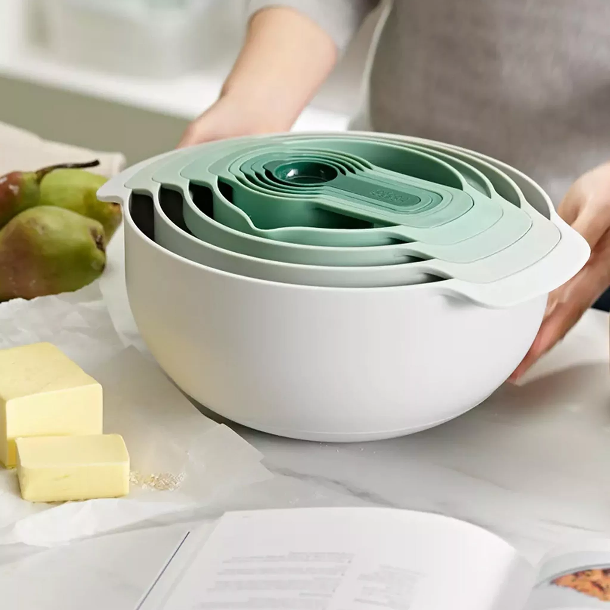 Stacked measuring bowls in white and green with some pears and block of butter by the side, plus a cookbook