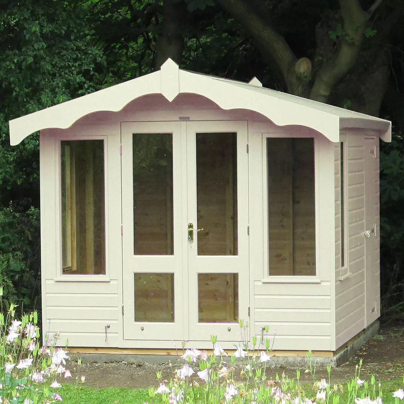 White timber garden summerhouse with double glazed doors and windows, set among trees and flowers.