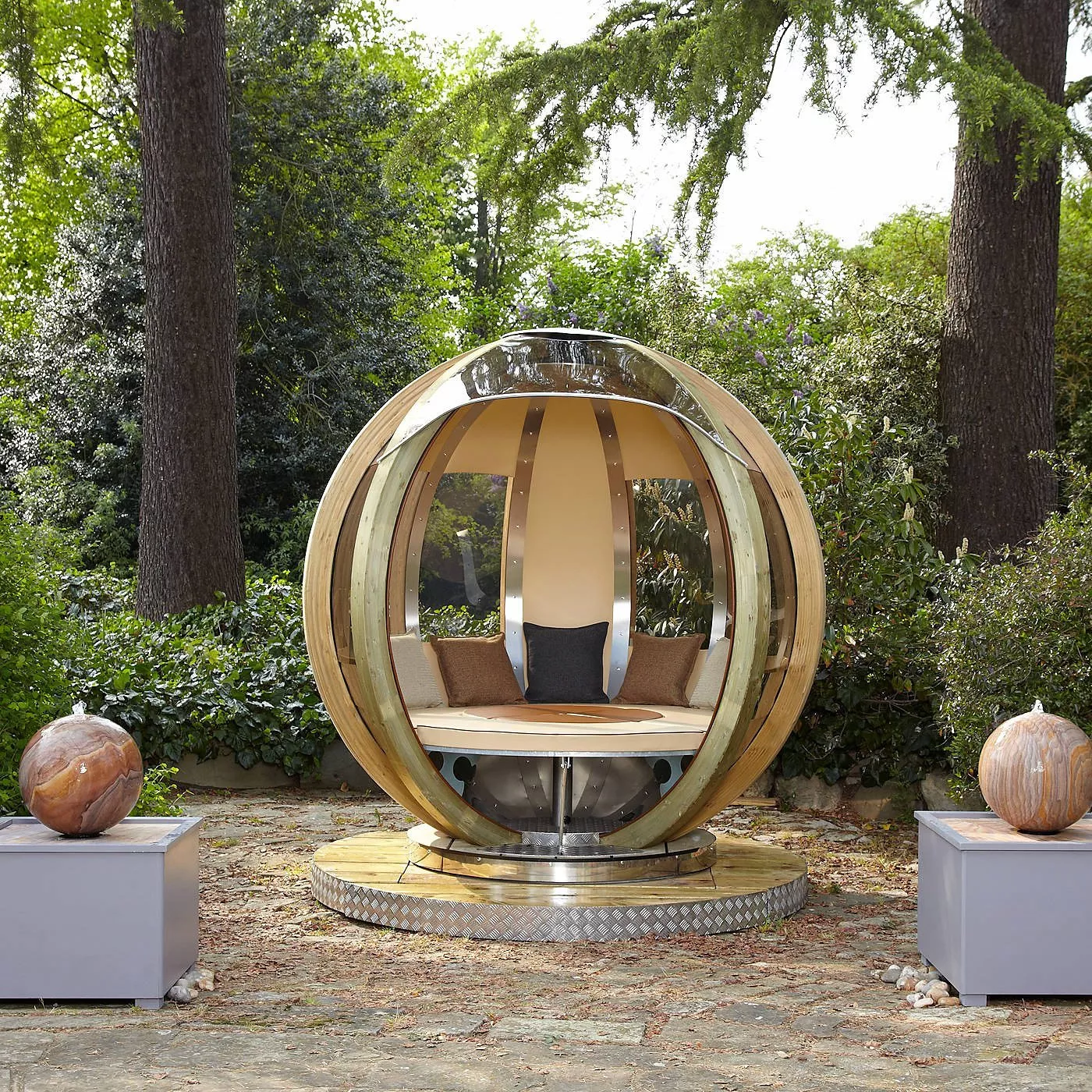 Modern spherical garden pod with cushioned seating, set in woodland with trees and stone patio outdoors