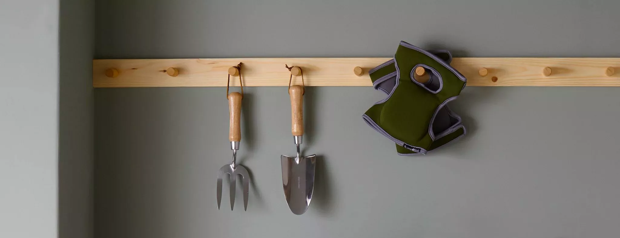 Wooden peg rail on grey wall holding garden hand fork, trowel and green dog harness for tidy storage
