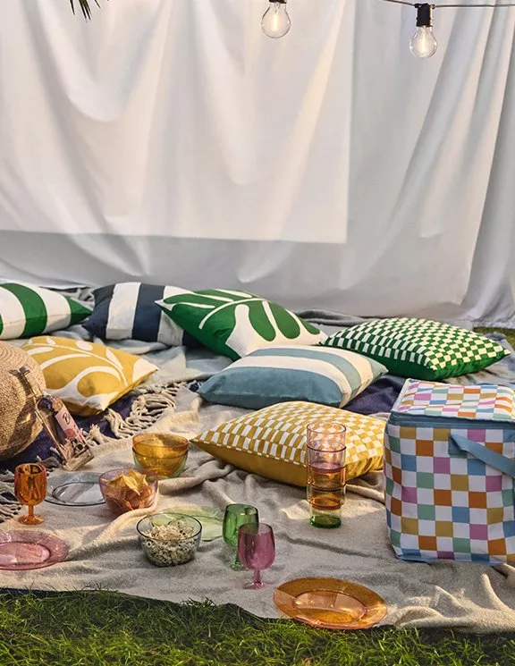 Outdoor picnic setup with colourful cushions, glassware and patterned cooler bag under string lights on grass