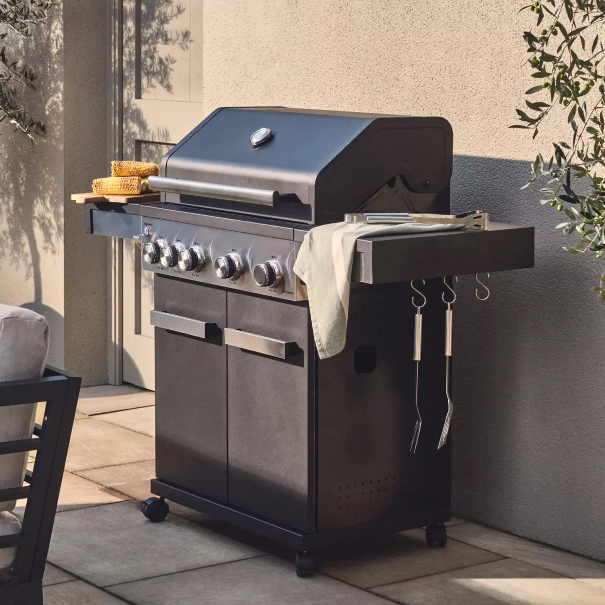 Modern outdoor gas barbecue grill with side shelf, utensils and corn cobs on a sunlit patio by a wall