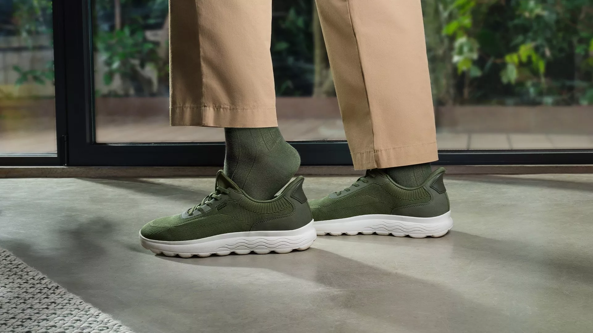 Person wearing olive green trainers, matching socks, and beige trousers on a concrete floor indoors.