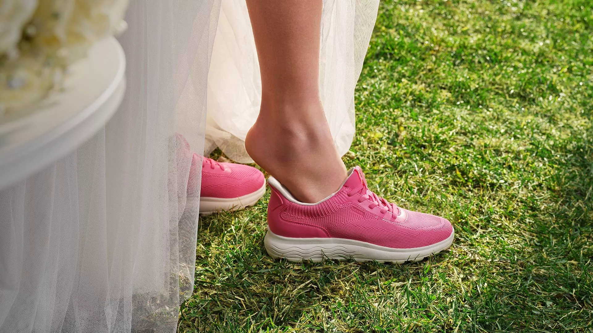 Bride in wedding dress wearing bright pink trainers on grass, showcasing stylish comfortable bridal footwear outdoors