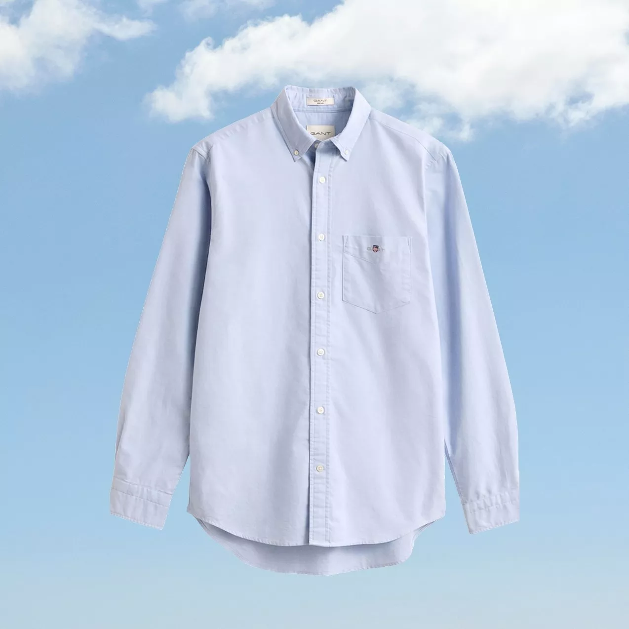 Light blue long-sleeve button-down Oxford shirt with chest pocket, front view on sky blue background with clouds