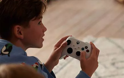 Child holding a white Xbox controller while playing video games indoors, close-up of hands and device