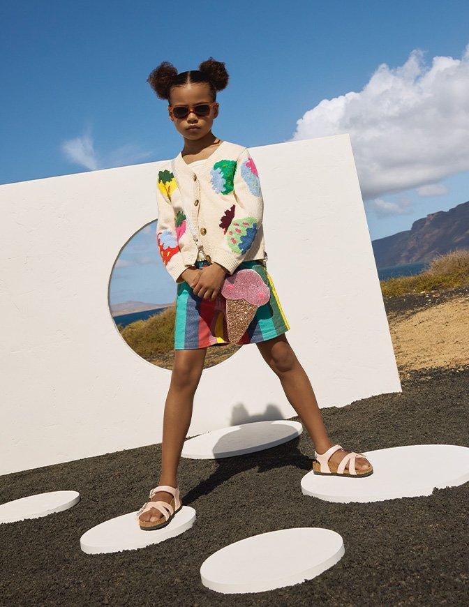Girl wearing colorful ice cream-themed outfit and sunglasses poses confidently outdoors against a white geometric backdrop