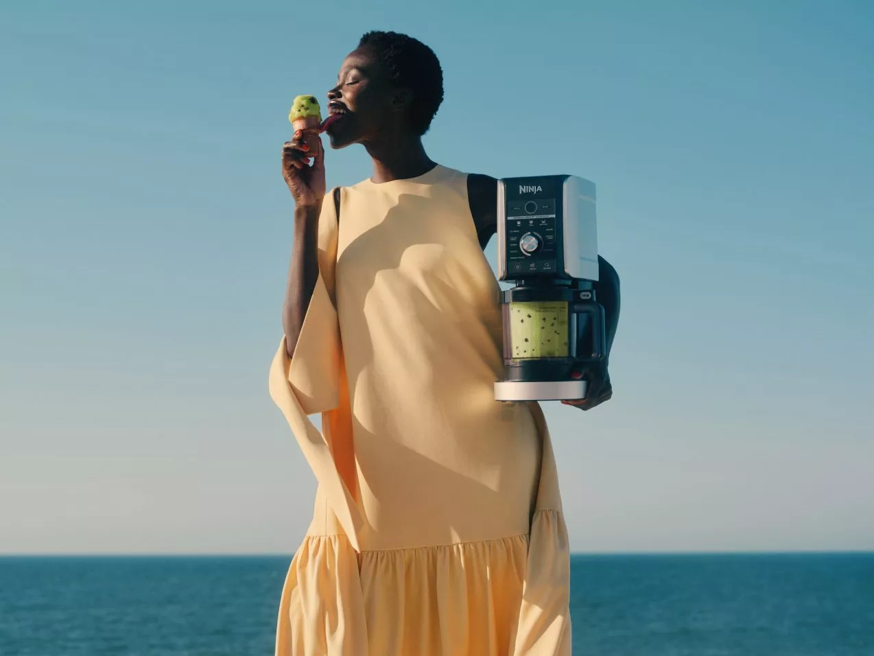 Woman by the sea holding a Ninja blender with green smoothie, enjoying an ice cream cone in sunshine