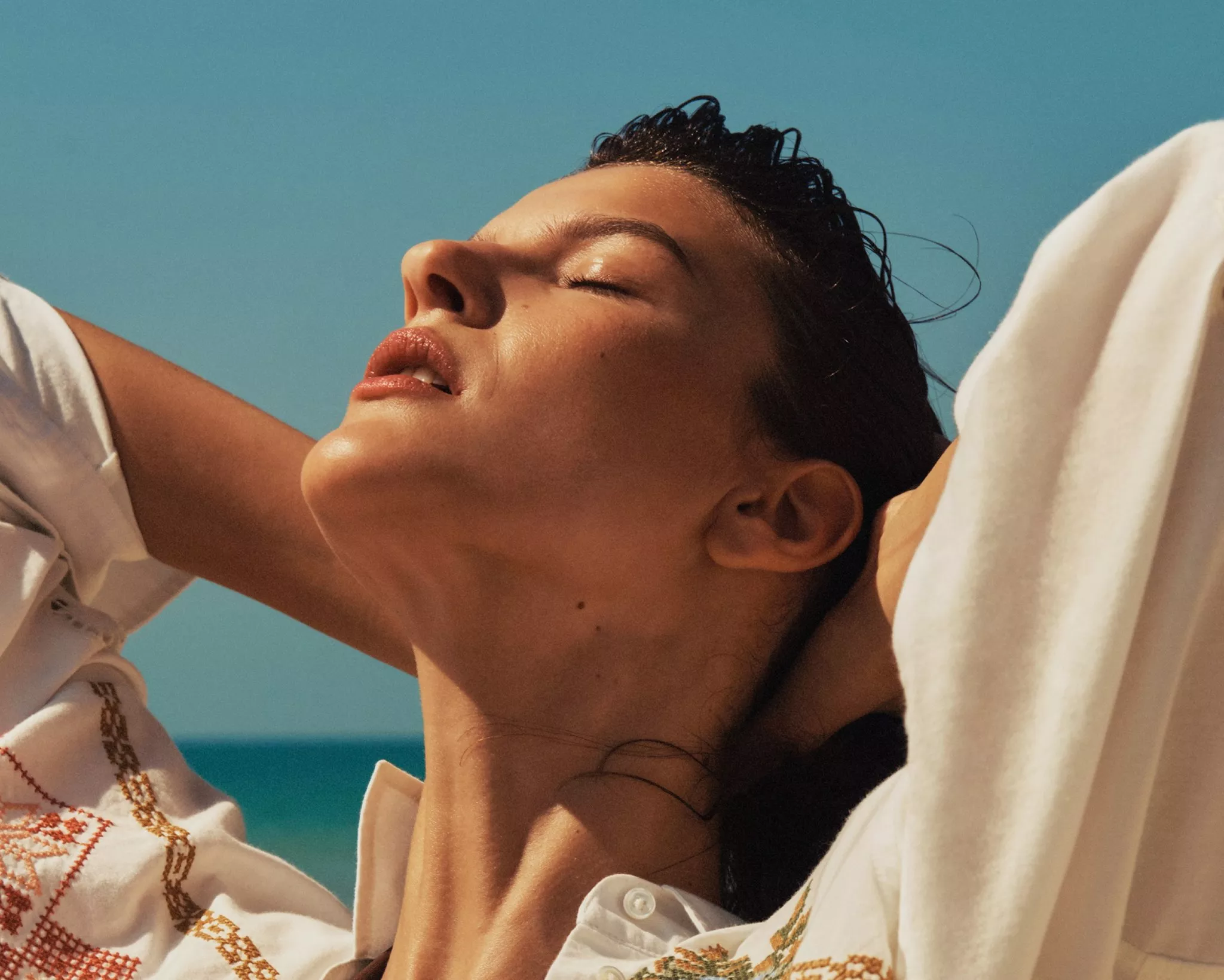 Sunlit woman relaxing with eyes closed by the sea, wearing white blouse, summer coastal lifestyle portrait