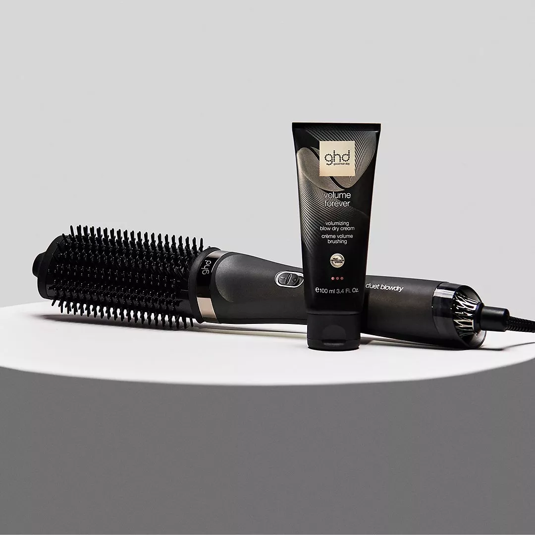 Black automatic hair curler on wooden table with comb, towel and perfume bottle, styled with eucalyptus leaves