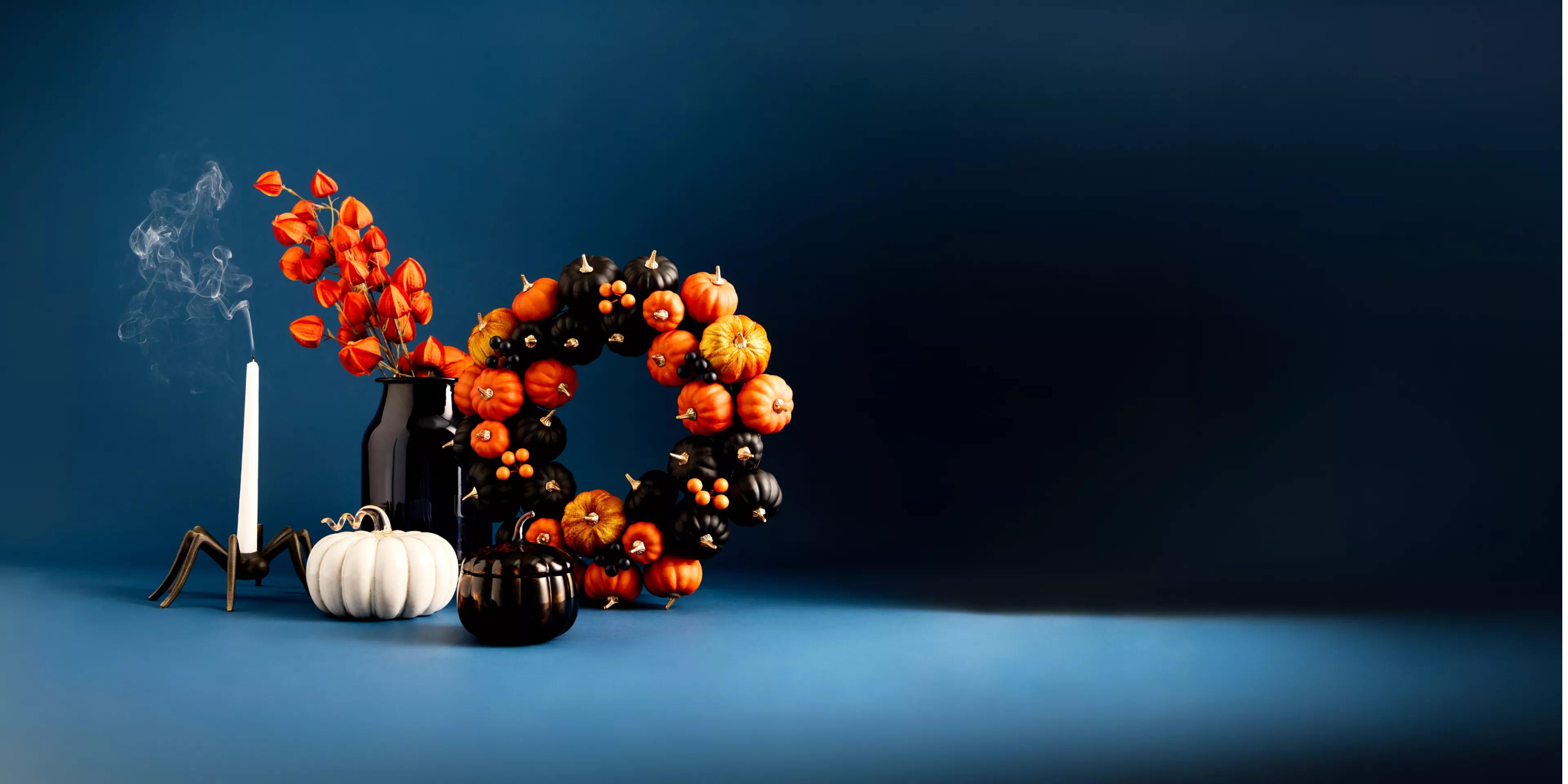 Halloween still life with pumpkin wreath, smoked candle, black vase and white pumpkin on blue background