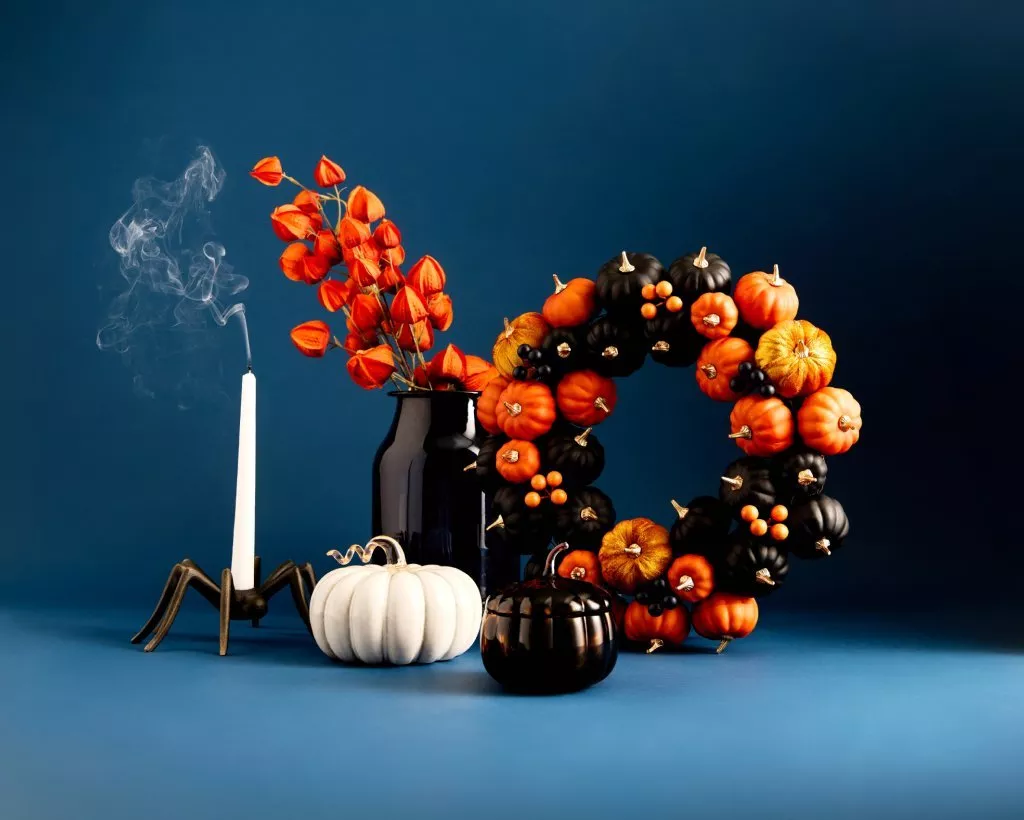 Autumn Halloween still life with pumpkin wreath, smoked candle, black vase and orange physalis on blue backdrop