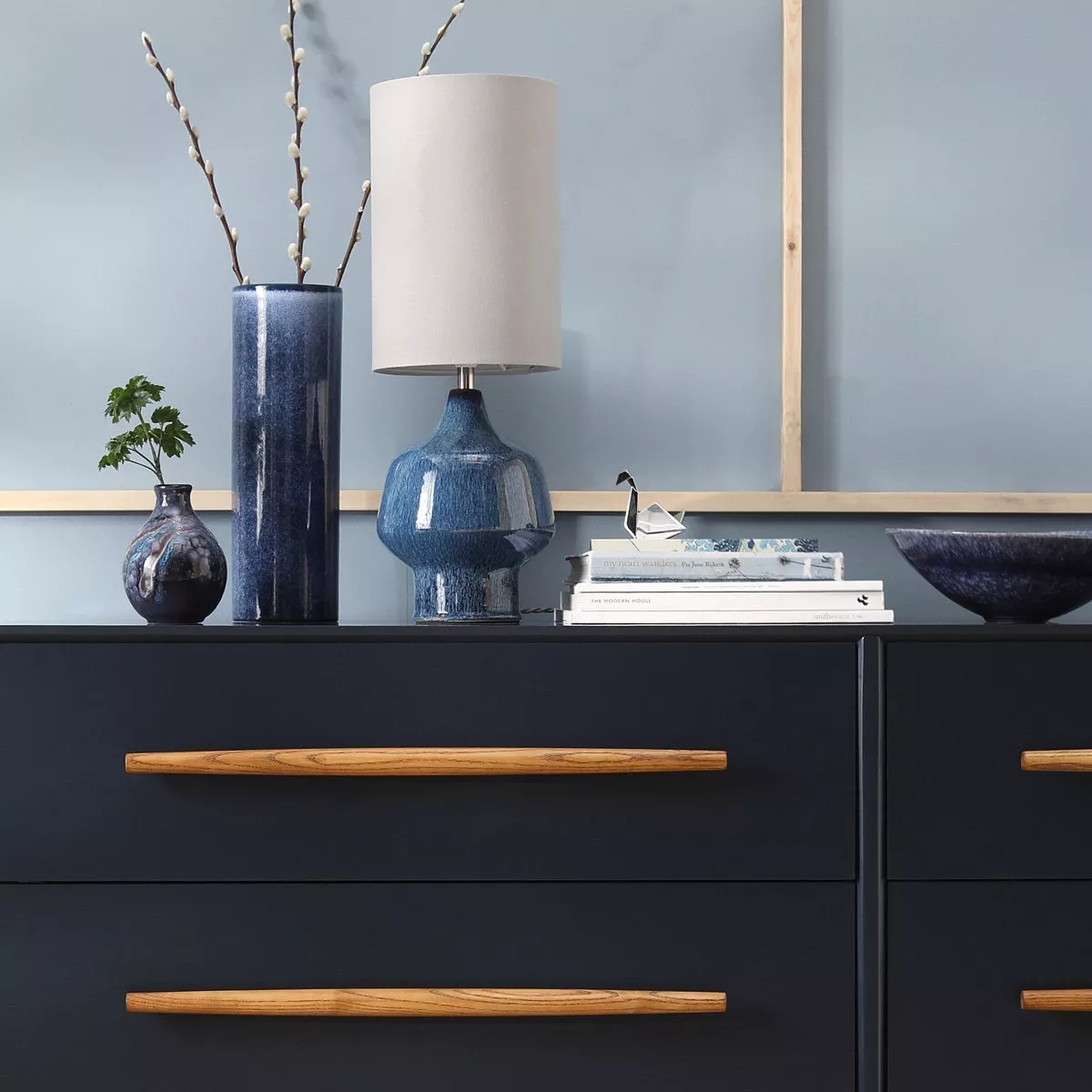 Modern sideboard styling with blue ceramic lamp, vases, books and bowl against a pale blue wall