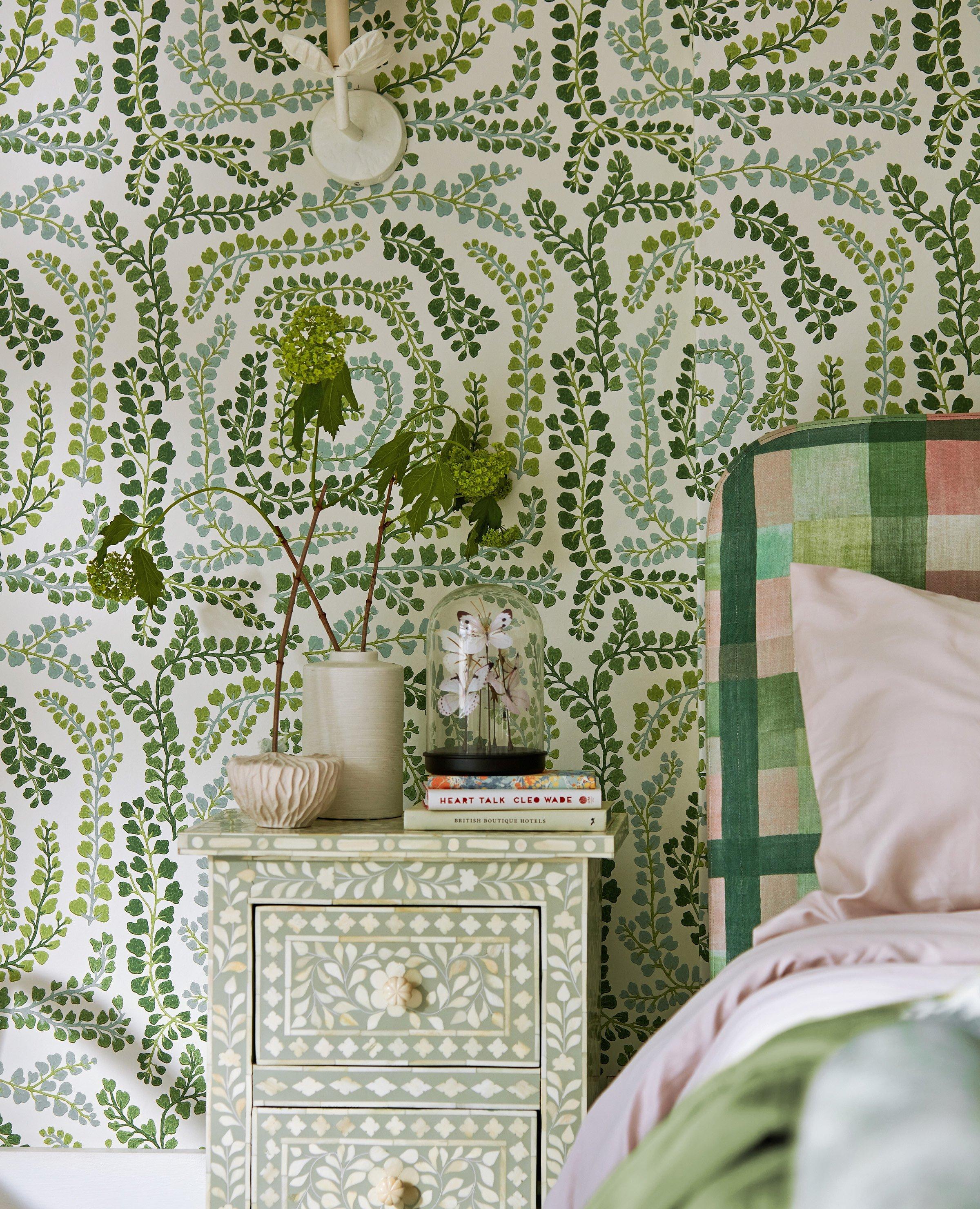 Harlequin green patterned wallpaper fashioned next to a bed with books and flowers on a bedside table