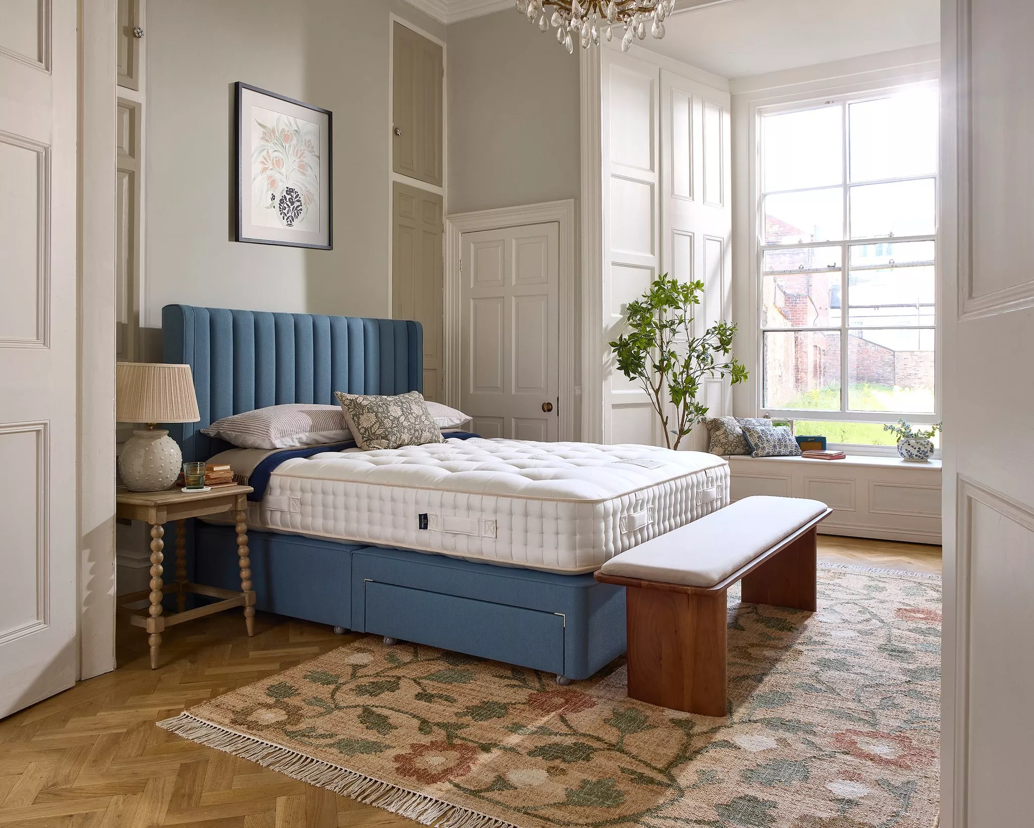 Bright bedroom with blue upholstered bed, white mattress, wooden bench, bay window and classic panelling