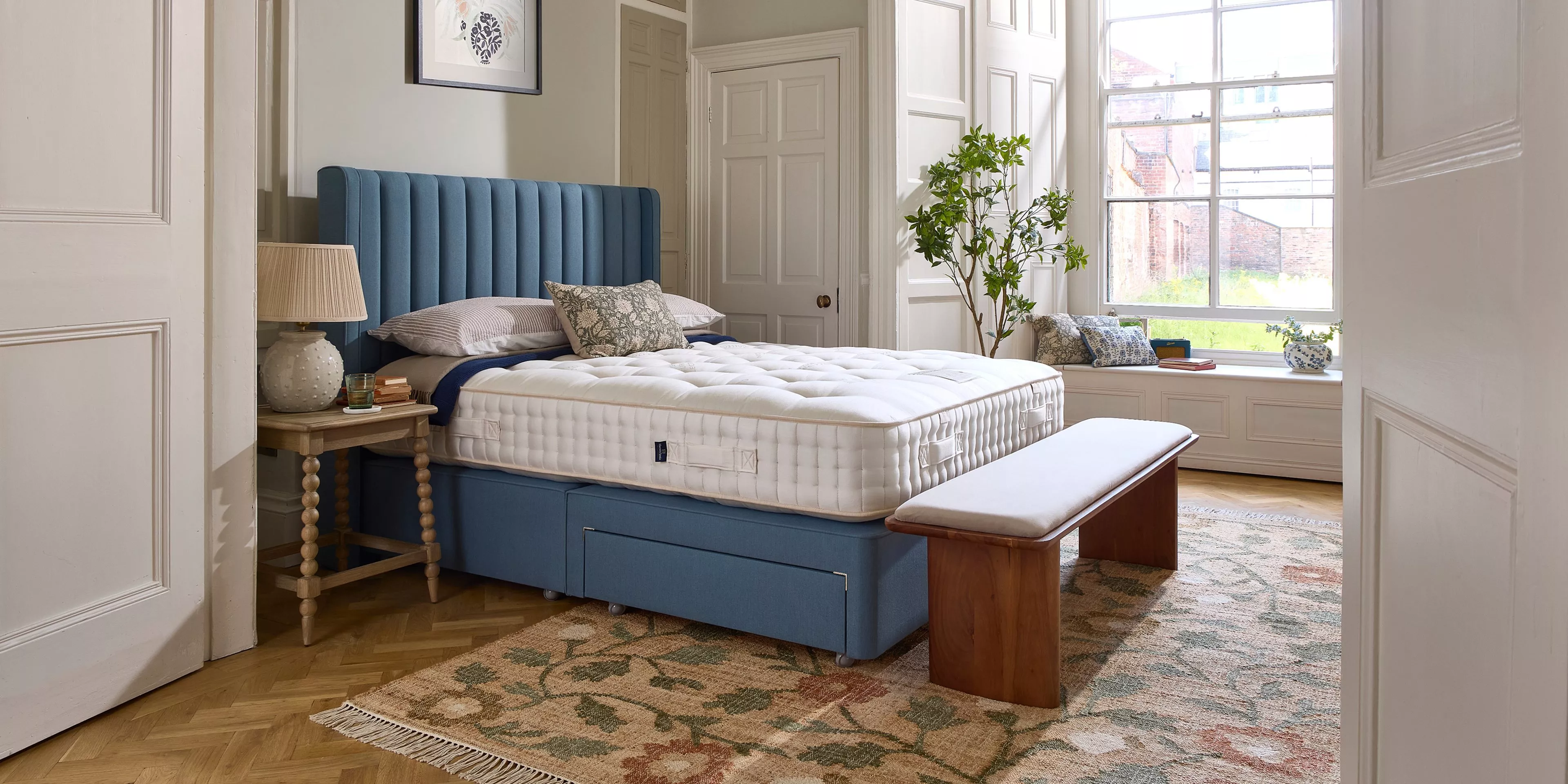 Cosy bedroom with blue upholstered bed, thick white mattress, wooden bedside table and floral rug on parquet floor