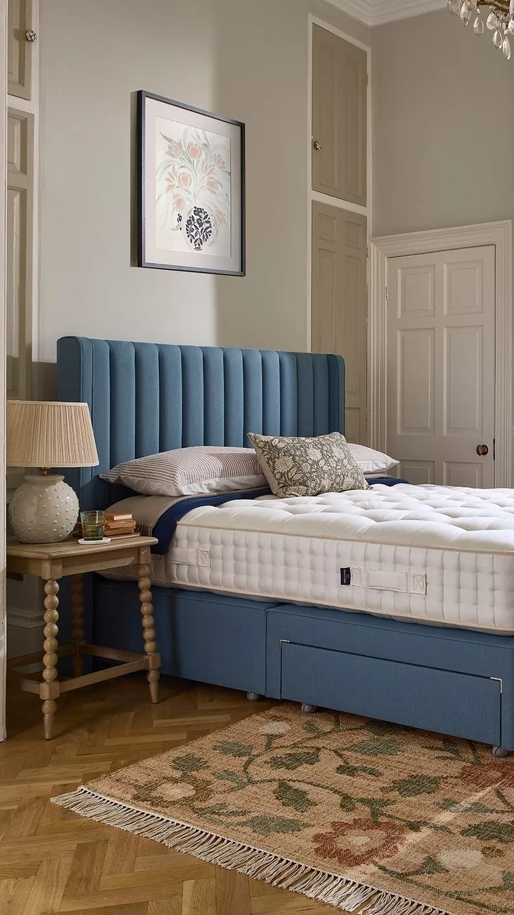 Cosy bedroom with blue upholstered bed, thick white mattress, wooden bedside table and floral rug on parquet floor