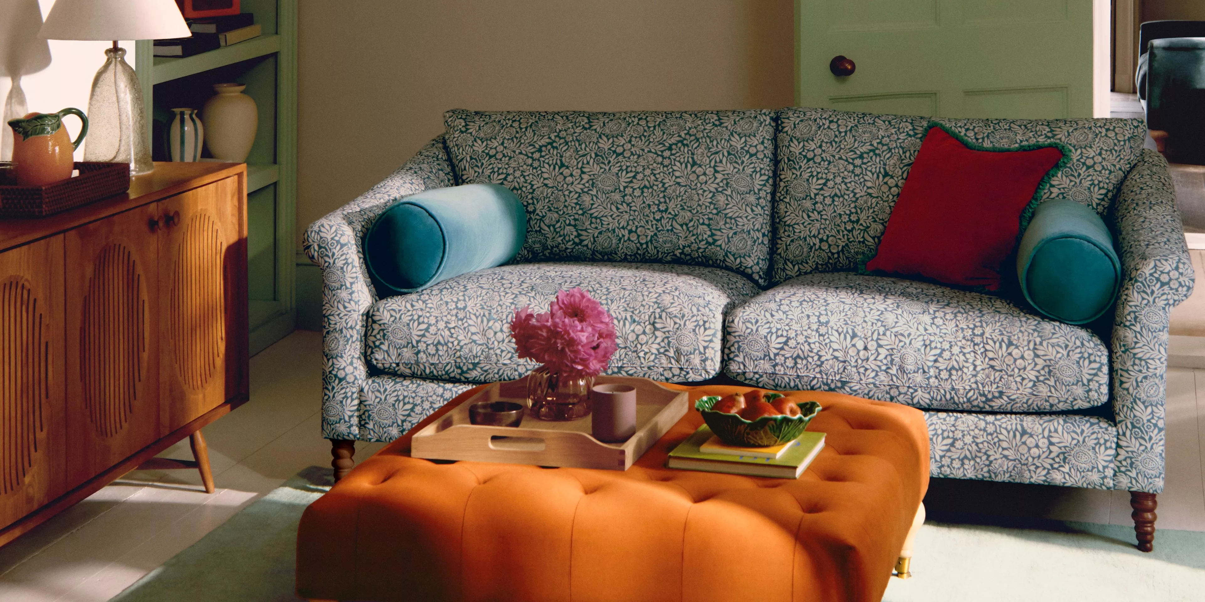 Stylish living room with a patterned blue sofa, vibrant cushions, wooden sideboard, and orange tufted ottoman.