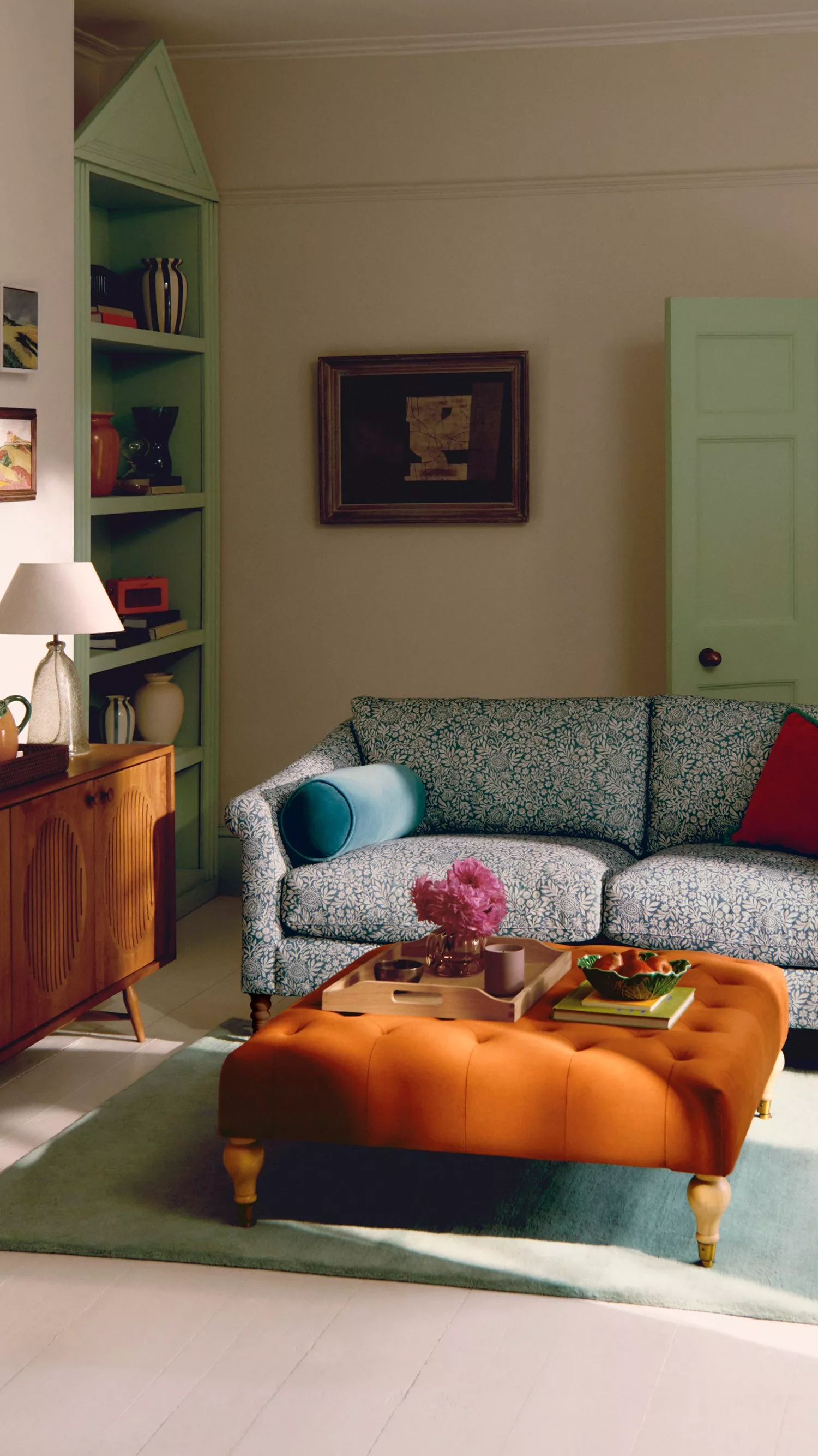 Stylish living room with a patterned blue sofa, orange ottoman, and a wooden sideboard. Decor includes a lamp and artwork.
