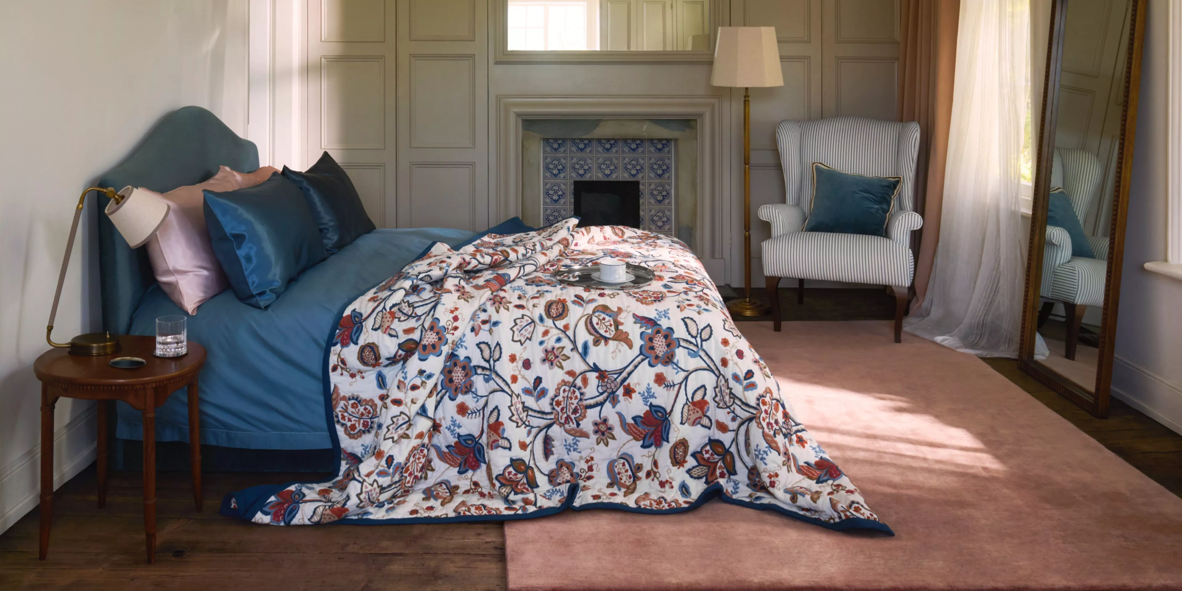 Cosy bedroom interior with blue upholstered bed, floral duvet, vintage mirror, tiled fireplace