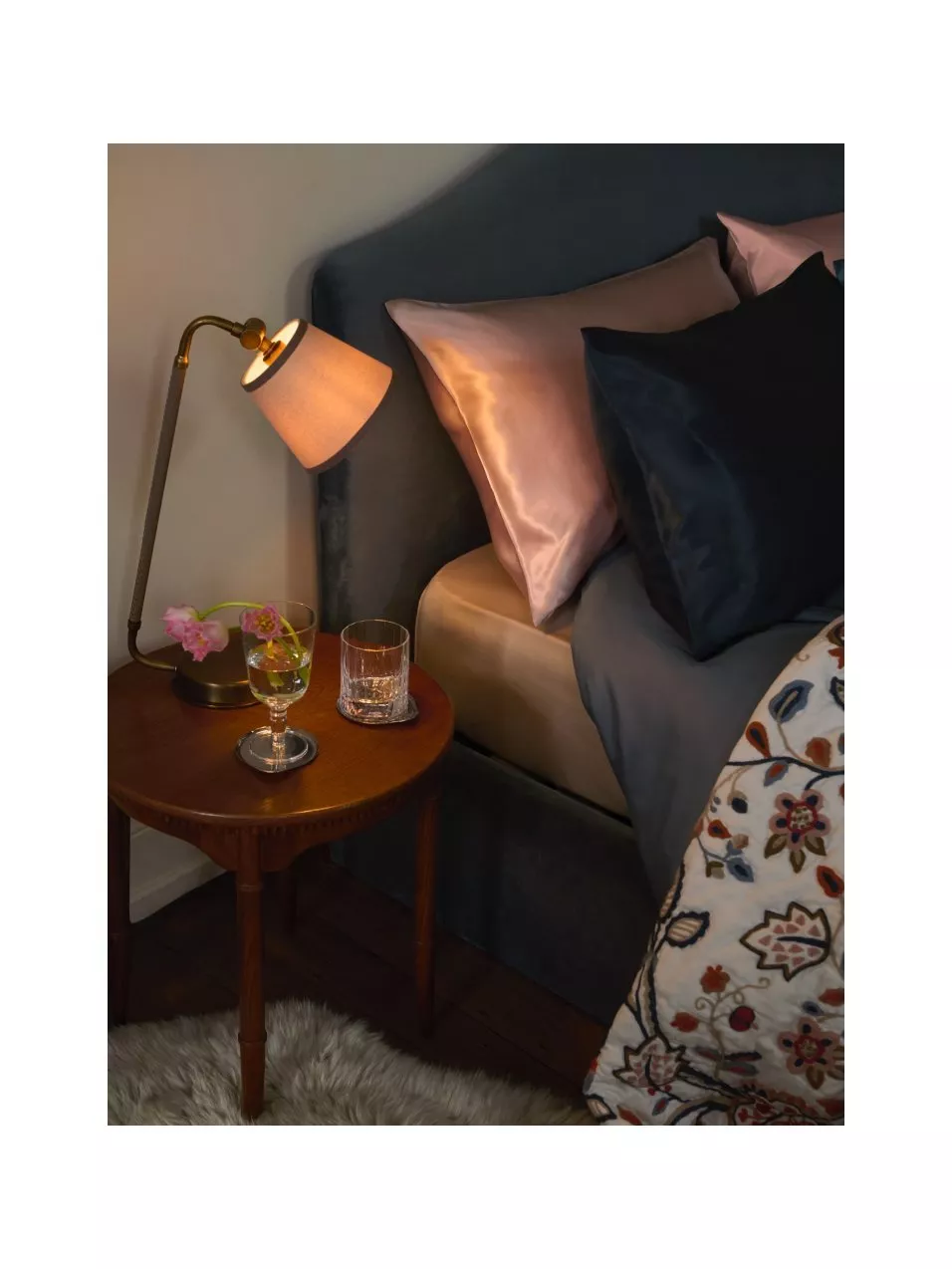 Cosy bedroom bedside table with brass reading lamp, beside blue upholstered headboard, pillows and floral bed spread
