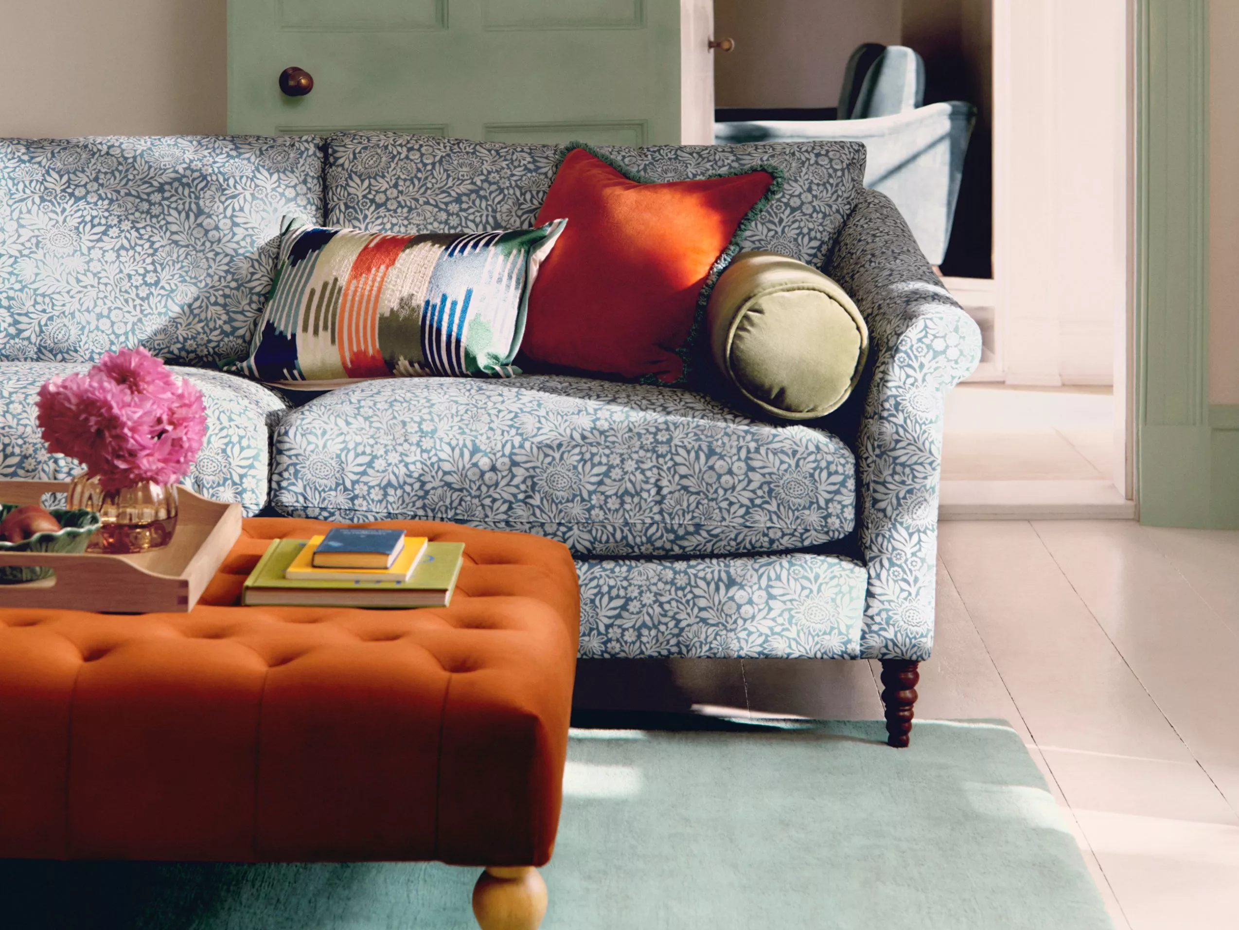 Cosy living room with a blue floral sofa, colourful cushions, an orange ottoman, and a soft pastel green rug.