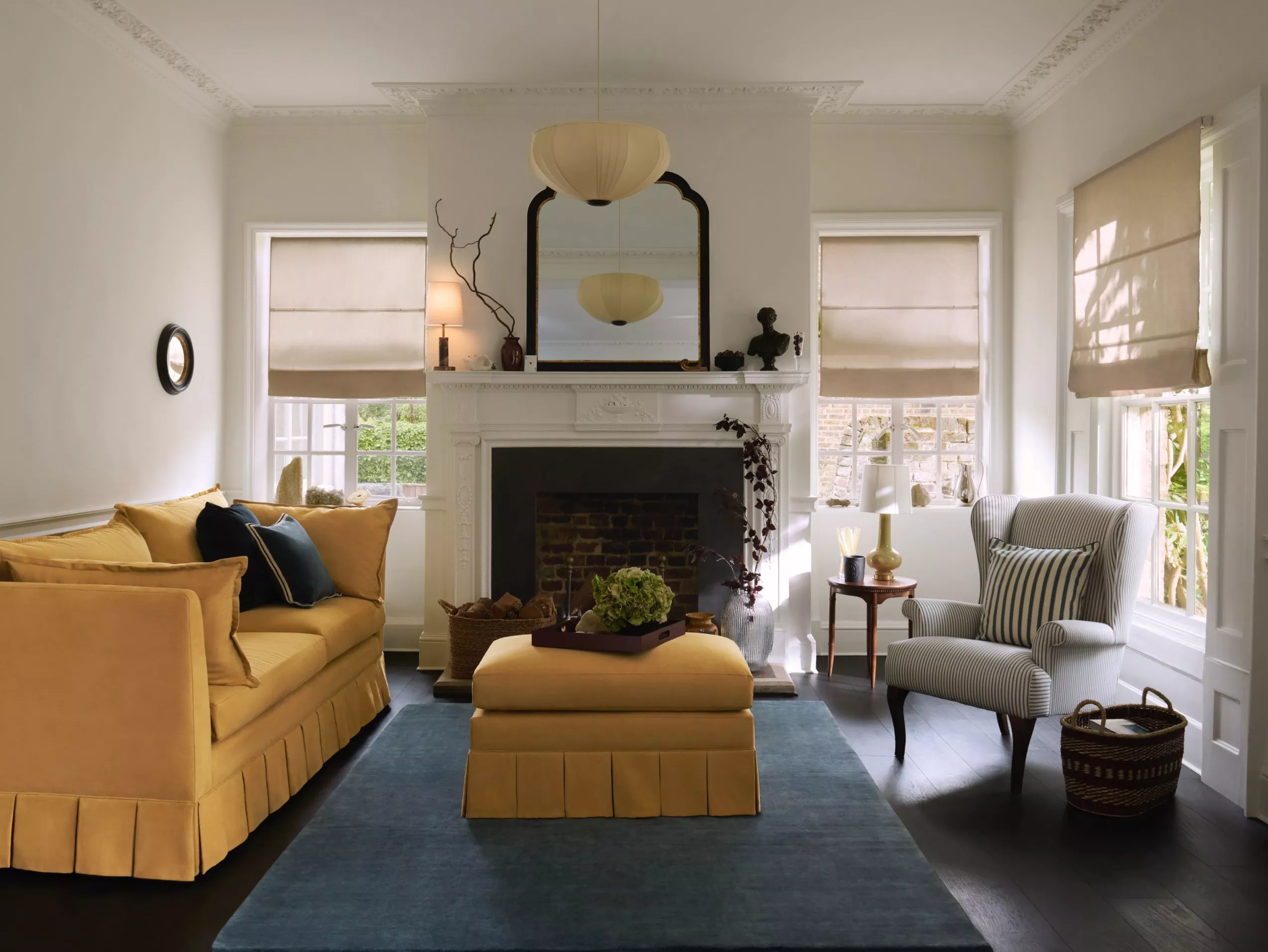Bright living room with yellow sofa, striped armchair, white fireplace, blue rug and roman blinds by windows