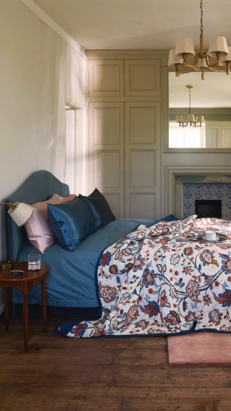 Cosy bedroom interior with blue upholstered bed, floral duvet, vintage mirror, tiled fireplace and brass chandelier.