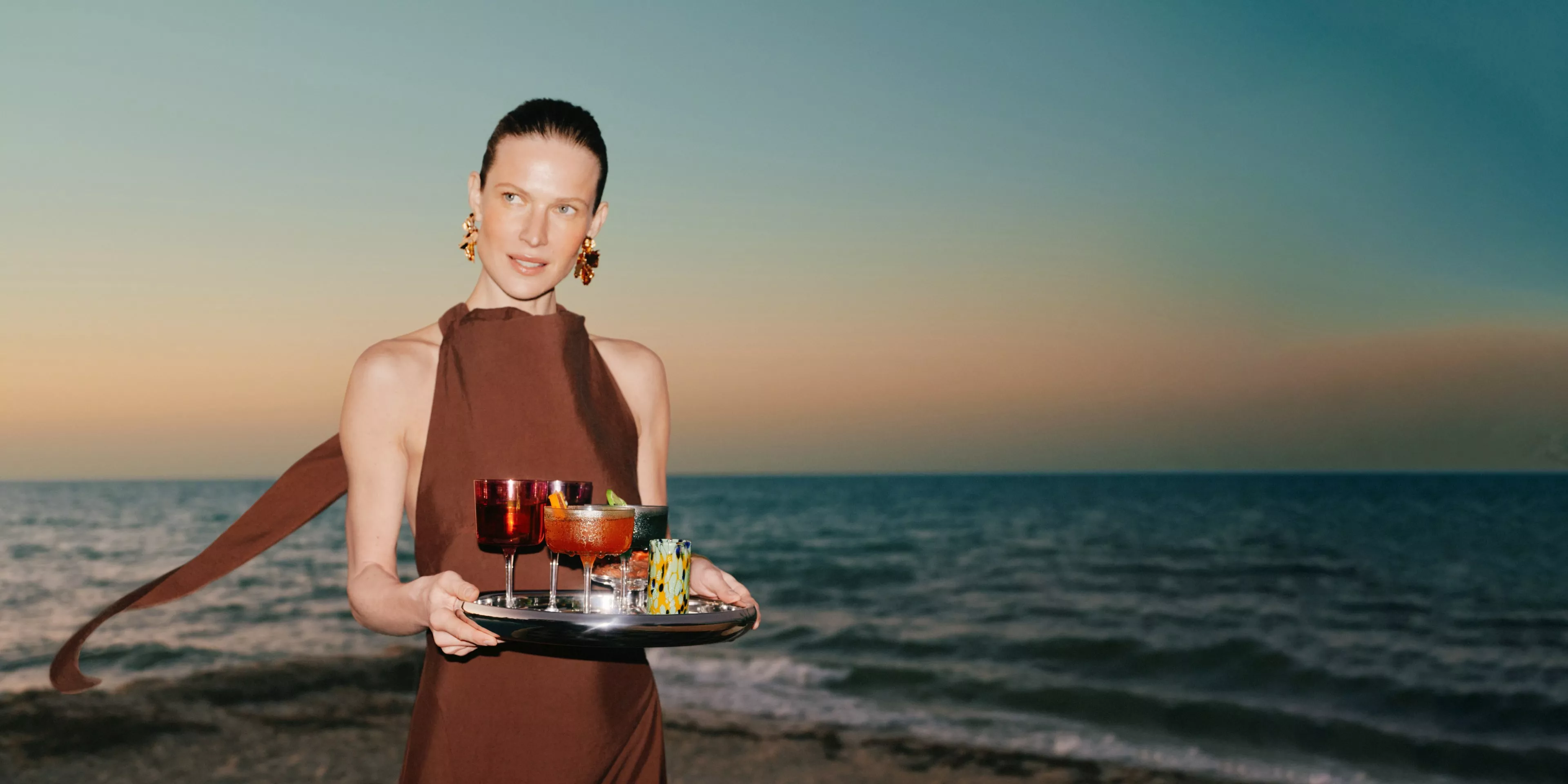 Stylish woman holding a tray of colourful cocktails on a beach at sunset, with sea and horizon backdrop