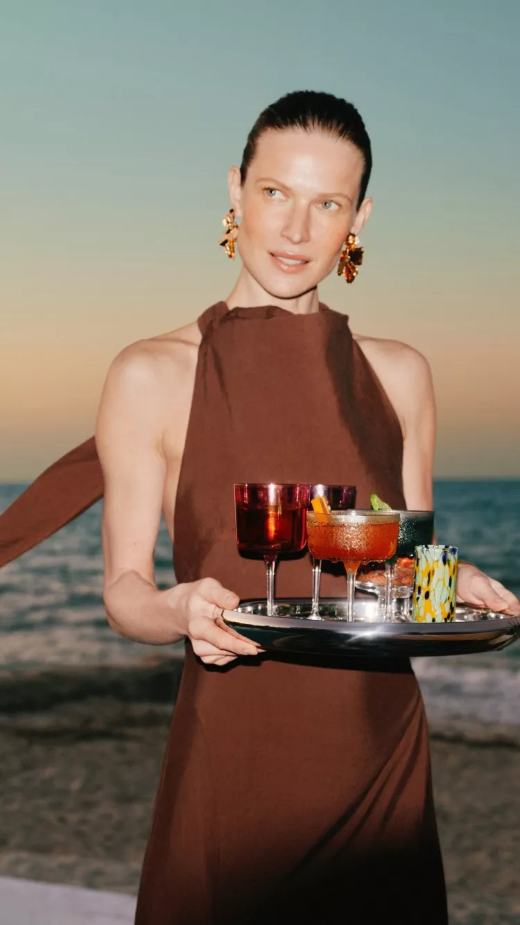 Woman in brown dress holding cocktail tray at seaside sunset, luxury beach bar drinks and glassware