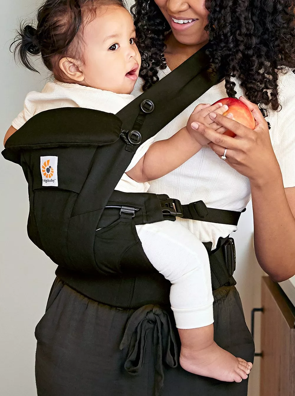 Woman carrying baby in a black ergonomic carrier, holding an apple, smiling. Baby dressed in white.