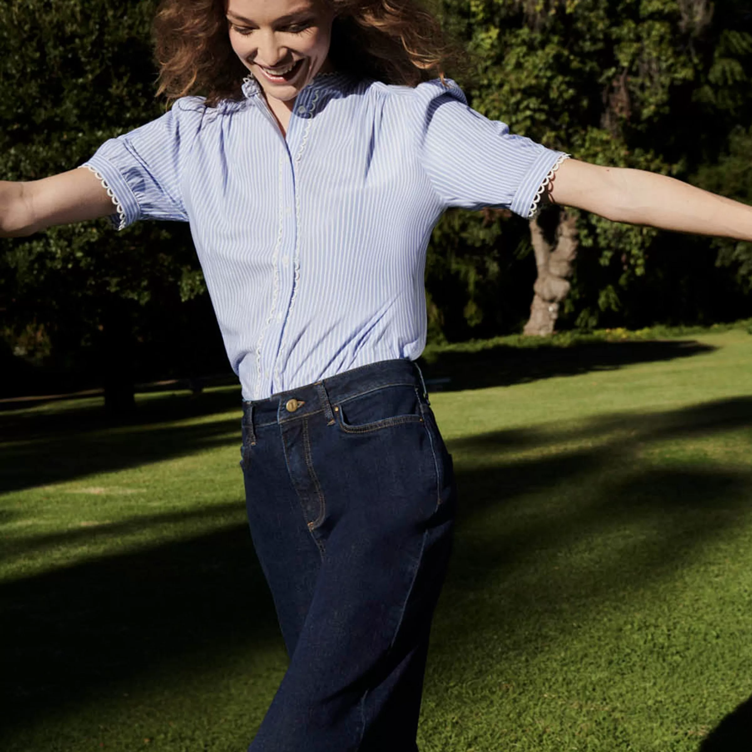 Smiling woman in blue striped blouse and dark denim jeans, arms outstretched on a sunny park lawn outdoors