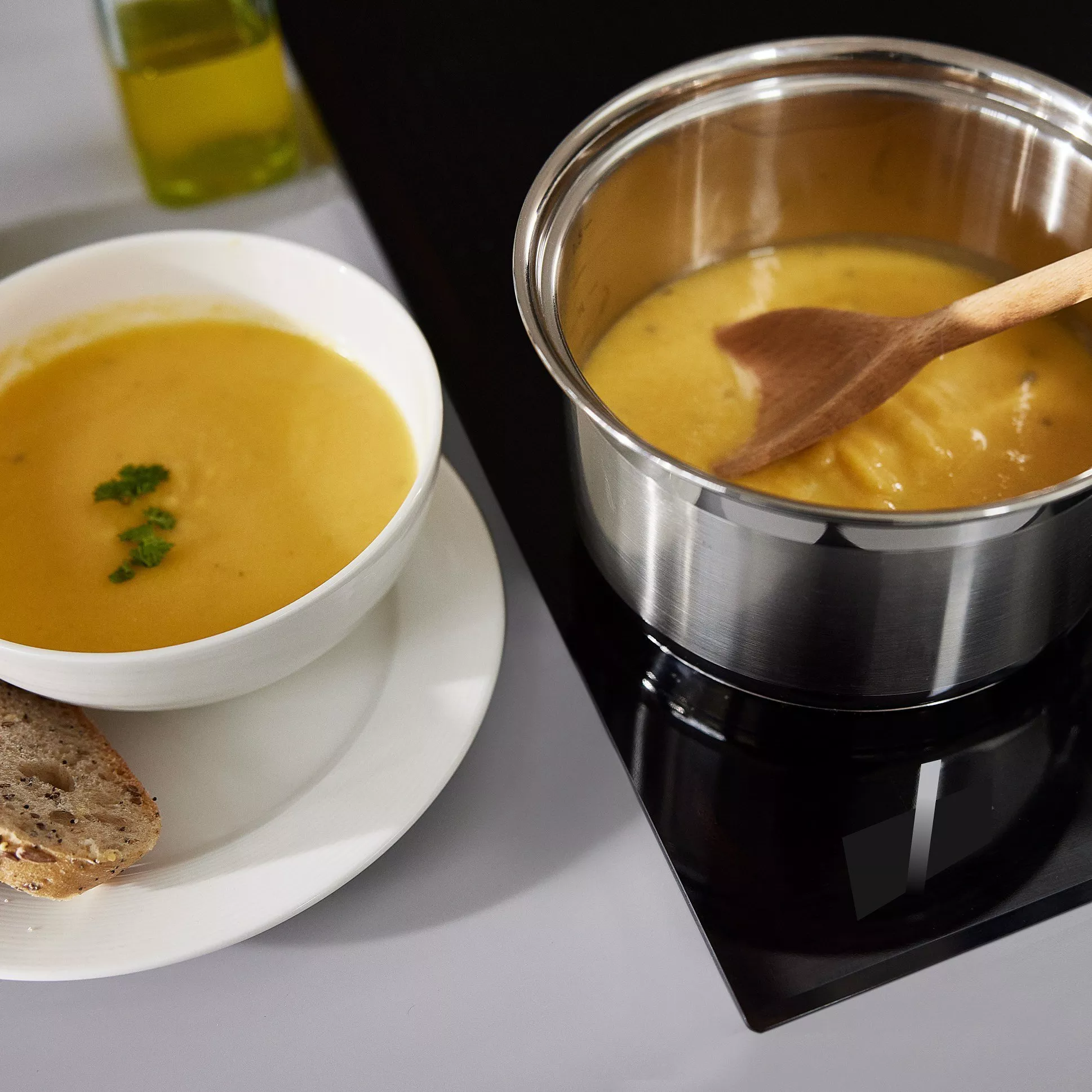 A bowl of creamy soup garnished with herbs next to a pot of soup on a stove, accompanied by crusty bread.
