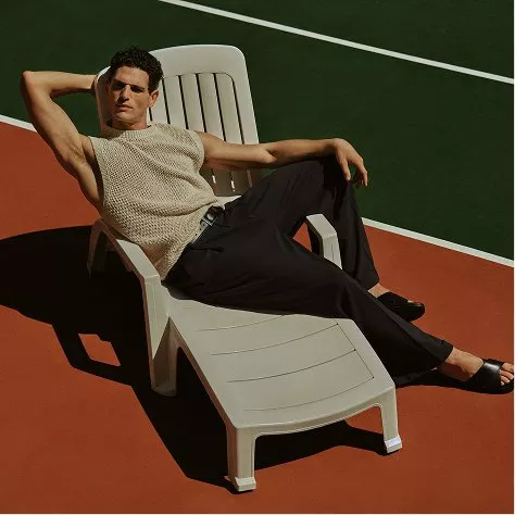 Man lounging on white sun lounger on outdoor tennis court, wearing sleeveless knit top and black trousers