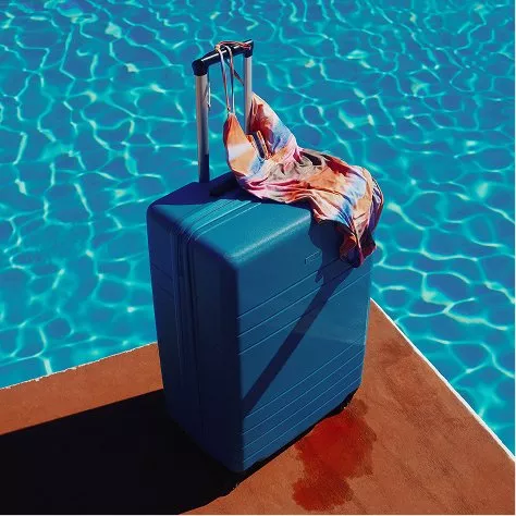 Blue hard-shell suitcase by a sunlit swimming pool, with colourful bikini draped over handle, summer travel vibe