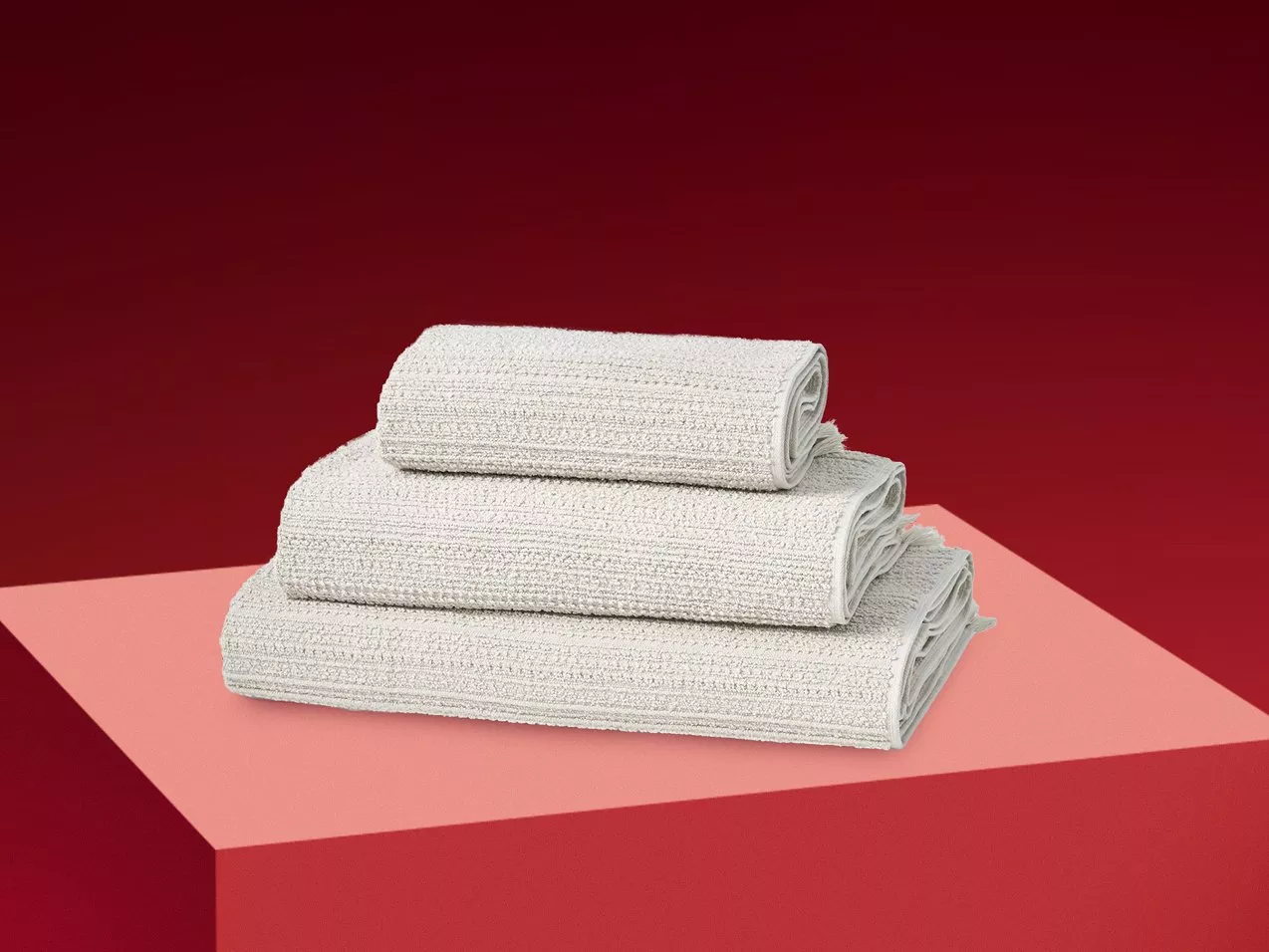 Stack of folded grey cotton towels on a pink pedestal against a deep red background, minimalist product shot